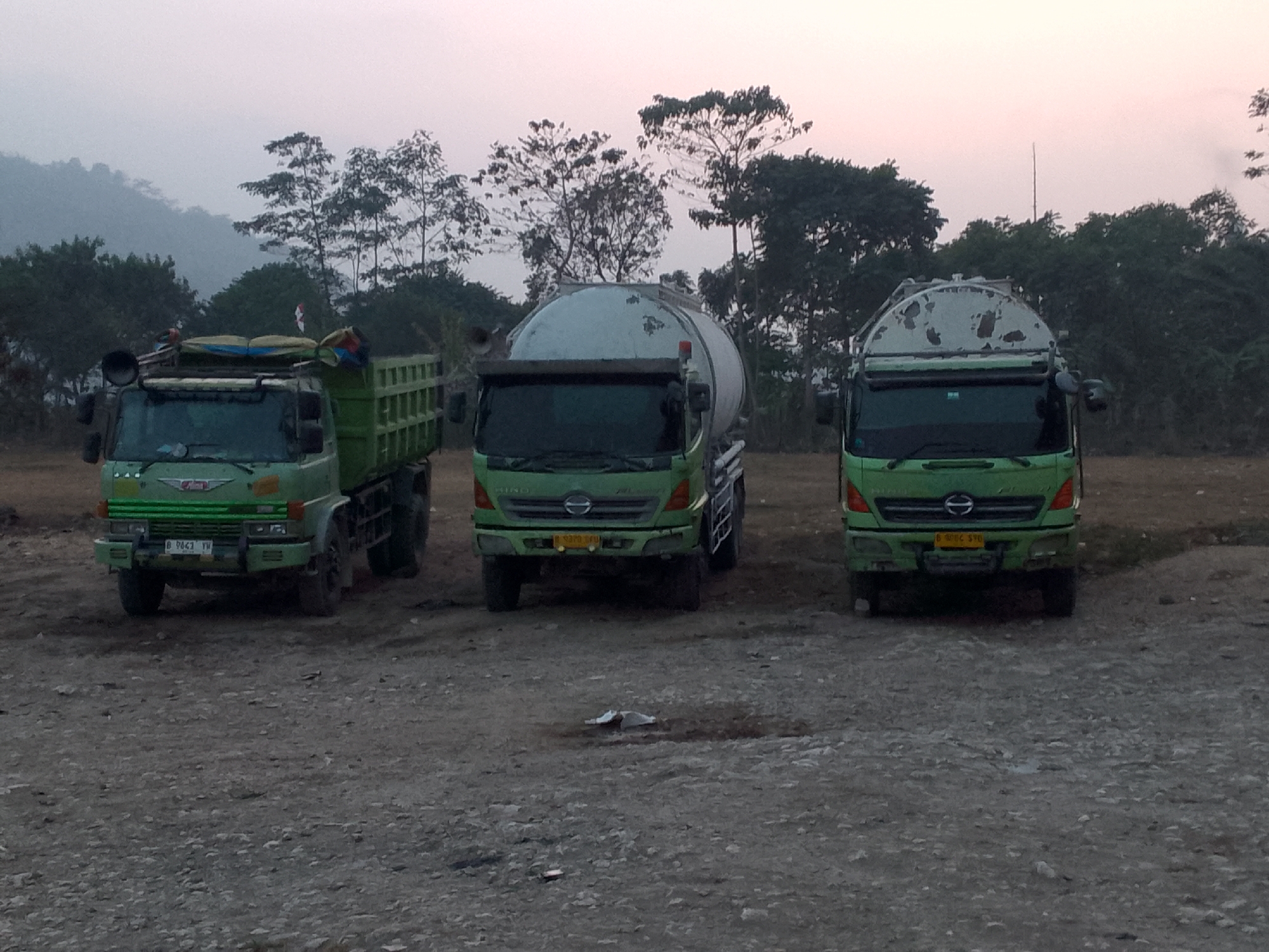 In Gunungmasigit, Indonesia, three green trucks are parked on a gravel lot. The truck on the left is a dump truck, loaded with cargo. The two trucks in the center and to the right are tanker trucks. The sky is overcast, and the scene is likely at dusk or dawn, with a light fog or haze in the background, obscuring the trees and hills.