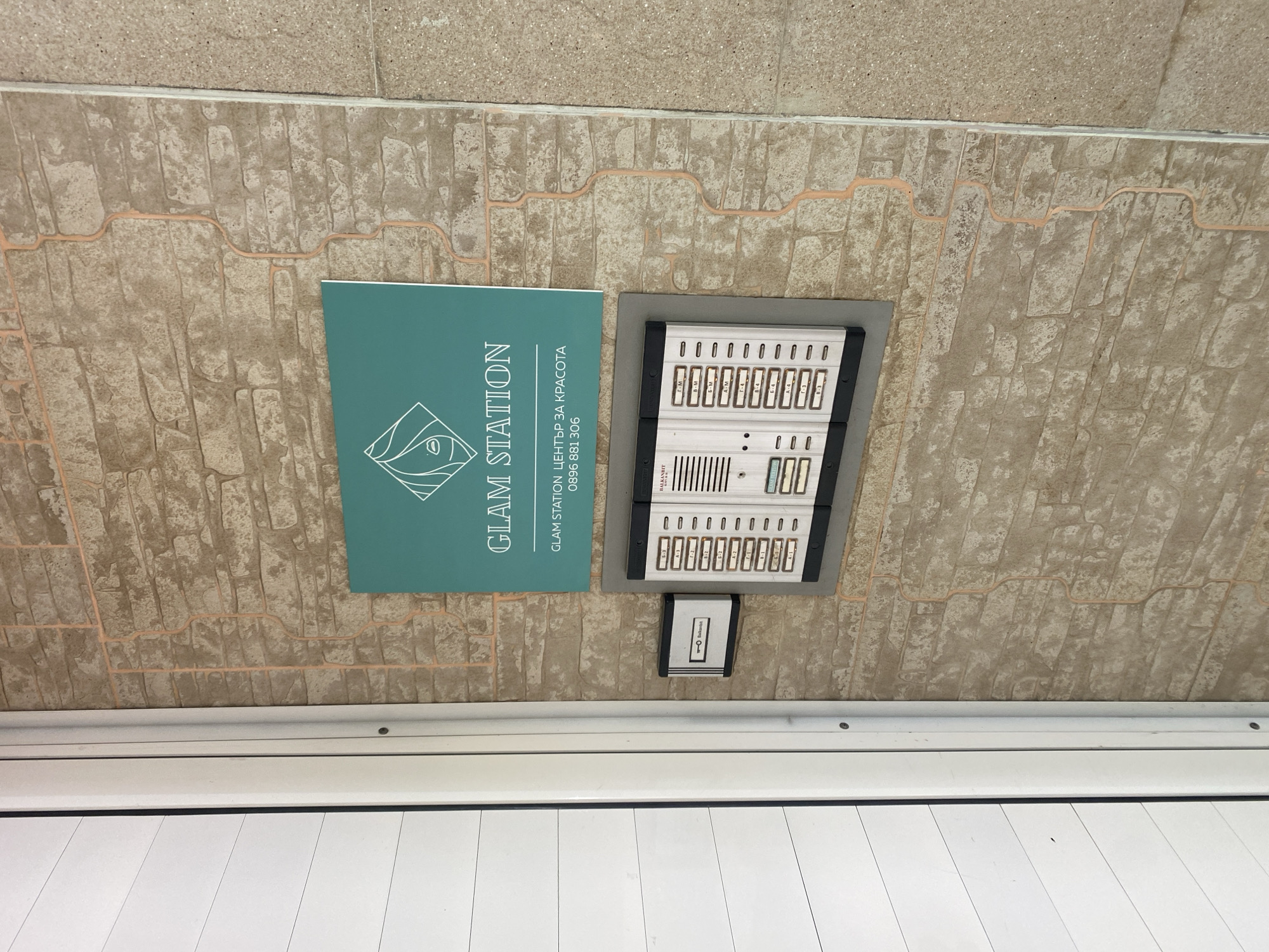 The image shows a sign and intercom panel attached to a wall. The sign, in teal, displays the words "GLAM STATION" vertically, accompanied by the text "GLAM STATION ЦЕНТЪР ЗА КРАСОТА" and the phone number "0896 881 306". Next to the sign is an intercom panel with several buttons labeled in Cyrillic characters alongside numerical designations, such as "г-м," "B-M," "Б-М," "A-M", "А-1". The wall has a brick pattern with painted seams. Below the wall, there is a white horizontal element, likely part of a door or window frame. Based on the Cyrillic text, the image is likely taken in Bulgaria, specifically in Sofia, where the phone number is likely based.