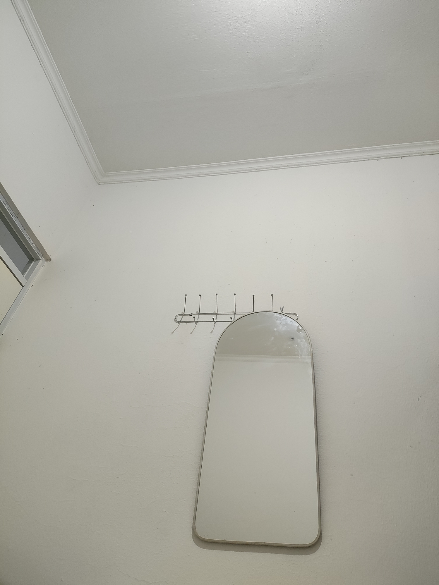 A light-colored wall is shown with a white ceiling that has decorative molding. A metal coat rack with multiple hooks is mounted on the wall. A curved, rectangular mirror with a thin metal frame hangs from the coat rack. To the left, a portion of a window with white trim is visible. The scene appears to be indoors, possibly in a room in Wanajaya, Indonesia, based on the provided context. No people or actions are depicted in the image.