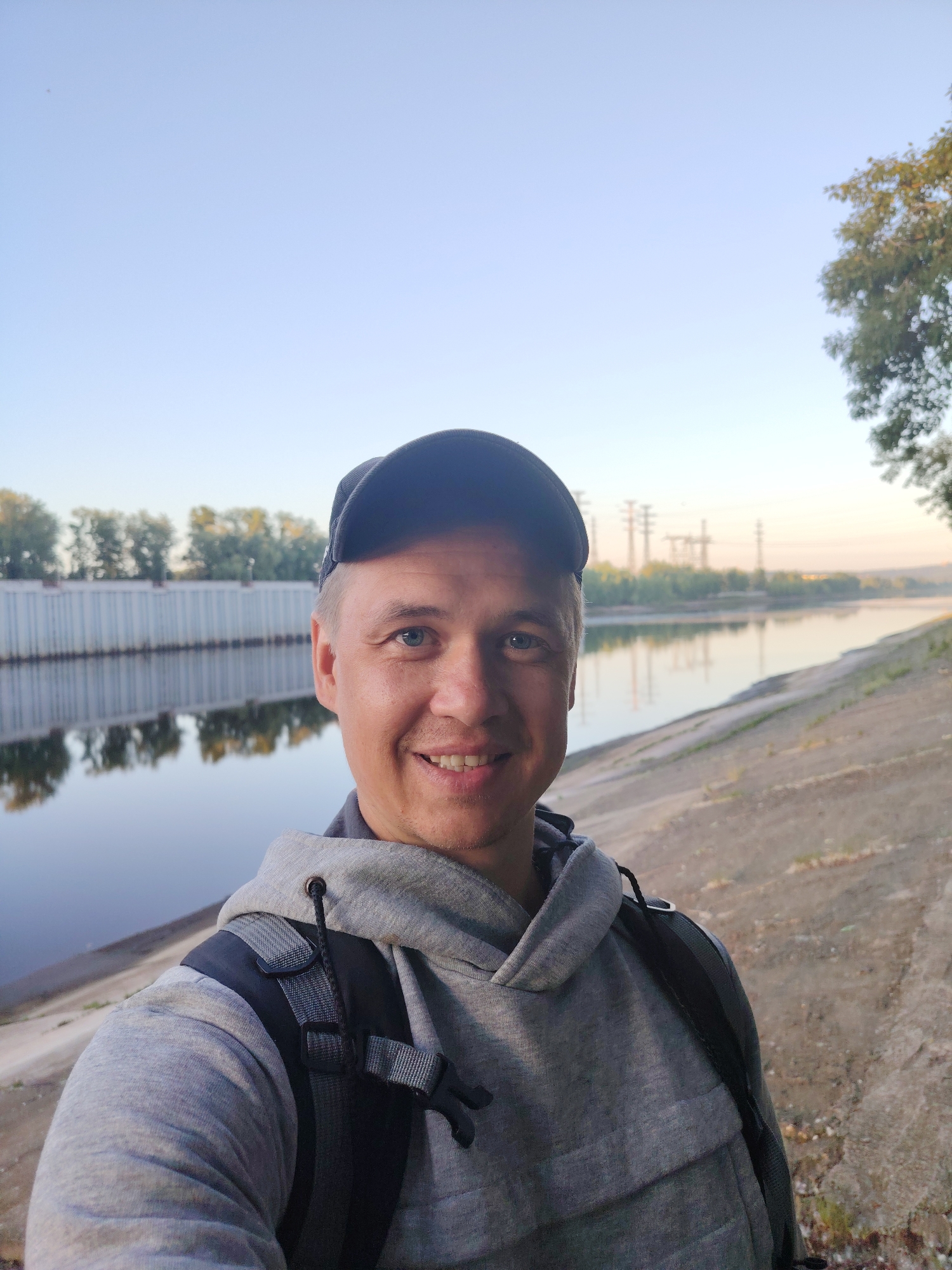 A selfie of a man wearing a gray hoodie, a black baseball cap, and a black backpack is taken outdoors. The man is smiling at the camera, with his right eye partially visible. He is standing on a sloped, dirt embankment next to a wide body of water, possibly a river or canal. The water reflects the sky and the opposite bank, which features a long, light-colored wall or structure lined with trees. In the distance, across the water, there are trees and industrial-looking towers against a clear, pale blue sky. The scene appears to be set in Chaikovsky, Russia, given the context. The lighting suggests it is either early morning or late afternoon.