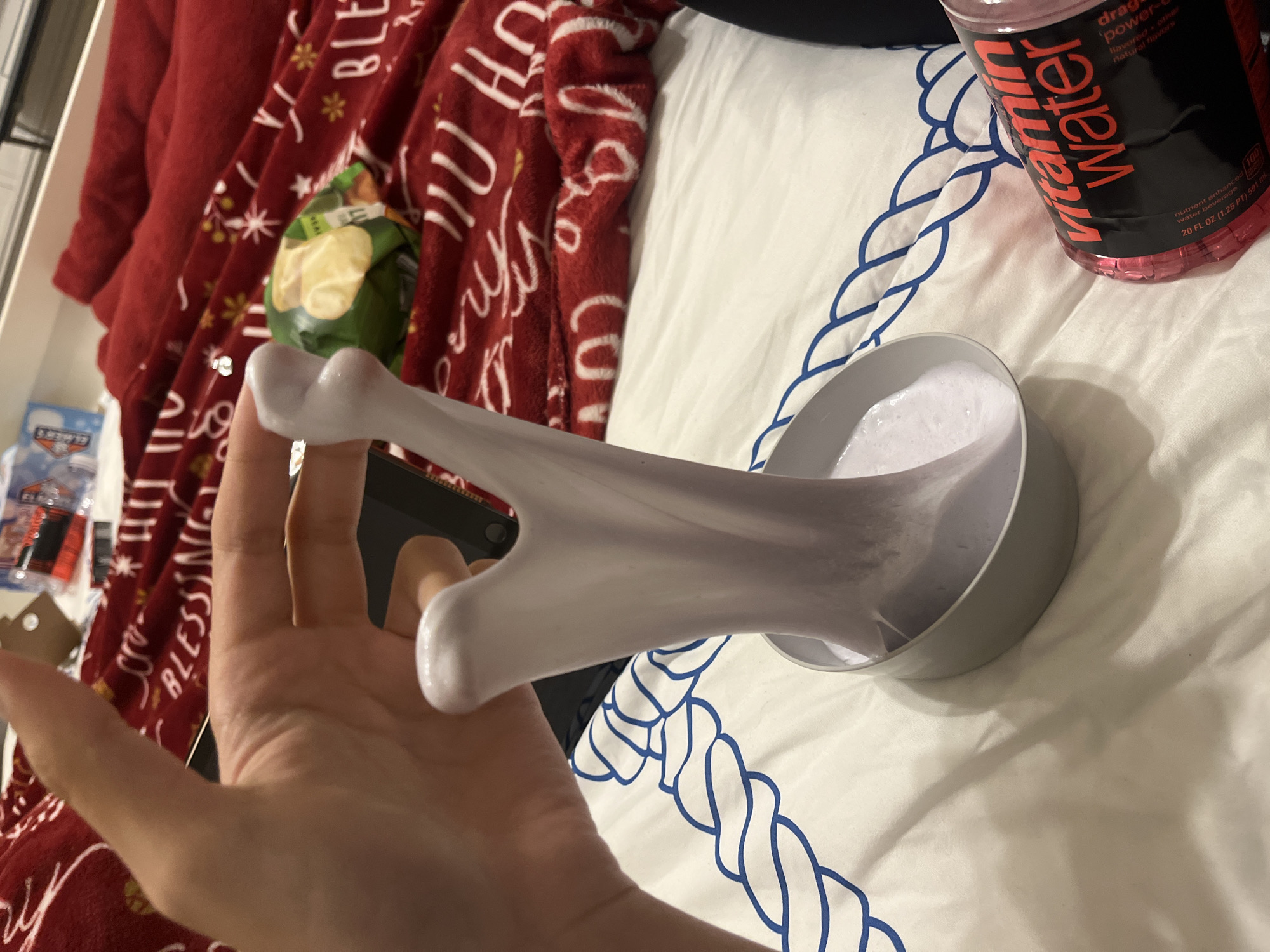 A hand stretches a white, putty-like substance from a grey bowl filled with the same material, against a backdrop of a white comforter with a blue rope pattern. A red blanket with holiday-themed white lettering and designs is partially visible. Next to the bowl is a bottle of Vitamin Water. A bag of potato chips and a tablet or small laptop are also visible in the scene. It is impossible to determine if the scene is in San Antonio, United States of America from this image alone.