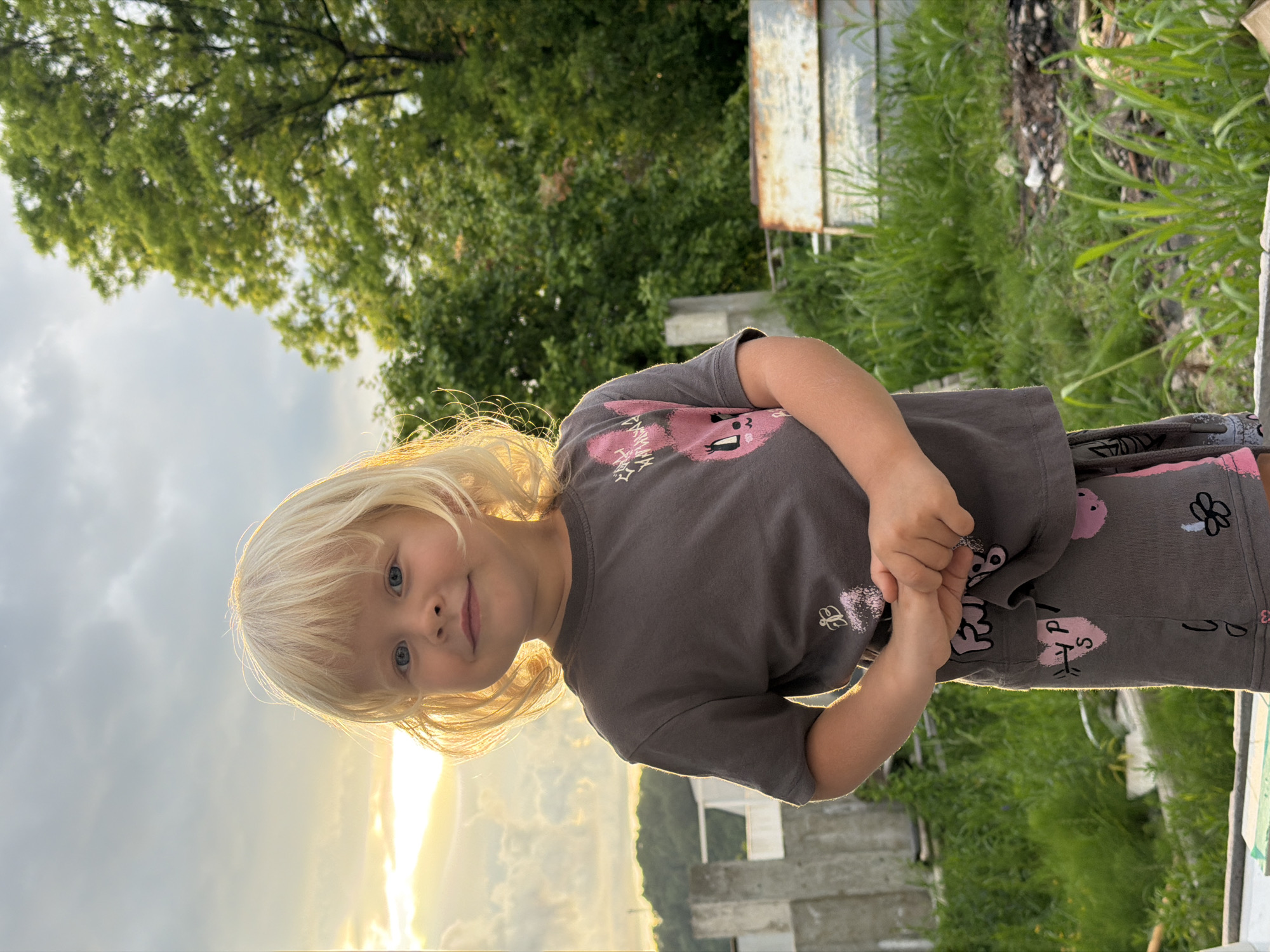 A young girl with blonde hair and blue eyes is shown outdoors, with the sun setting behind her, casting a warm glow. She is wearing a grey t-shirt with a pink design of a cartoon character and matching grey shorts with a similar pattern. Her hands are clasped together in front of her. The background includes lush green trees, tall grass, and some weathered wooden structures. The sky is cloudy, with rays of sunlight breaking through. The scene appears to be a rural or backyard setting, possibly in Baranovka, Russia, during the evening. The girl has a gentle smile, suggesting a calm and pleasant mood. There is no visible text in the image.
