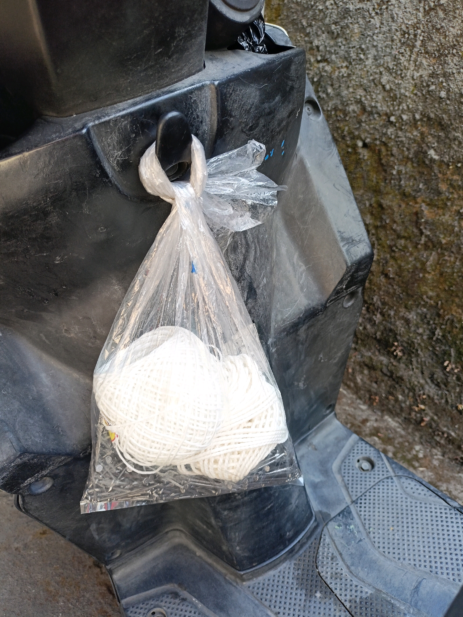 A plastic bag containing a large amount of white twine and a collection of nails is hanging from a black hook on the side of a black scooter. The scooter is parked next to a textured, rough-surfaced wall, possibly concrete or stone, with some moss or discoloration. The ground appears to be a mix of dirt and pavement. The image is taken from a slightly elevated, close-up perspective. The location is identified as Payaman, Indonesia.