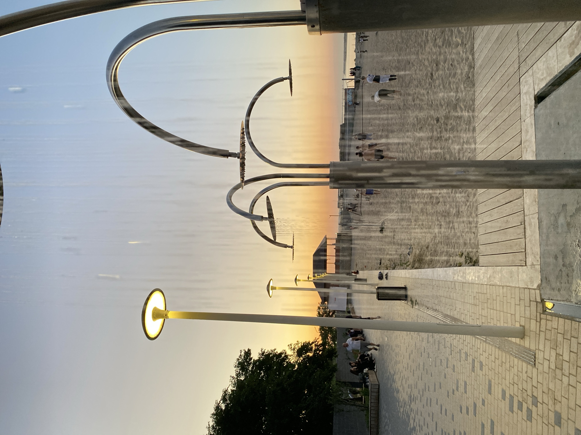 The image captures a sunset scene at a beachside promenade in Makhachkala, Russia. The sky is illuminated with warm hues of orange and yellow, indicating the late afternoon or early evening. Several people are scattered across the sandy beach and the paved walkway, appearing to be enjoying the serene atmosphere. Some individuals are seen walking or sitting, while a few appear to be children playing.
Dominating the foreground are elements of a water feature, likely a public spray park or fountain, with sleek, modern metallic structures and arcs. Water is visible, creating a shimmering effect that reflects the sunset. Along the walkway, modern lampposts with circular illuminated tops stand at intervals, casting a warm glow. The pathway is paved with light-colored bricks and has wooden decking sections. In the background, the beach stretches out, meeting the calm sea.
The overall mood is peaceful and relaxed, with people taking advantage of the pleasant weather and the scenic view of the sunset over the water. The presence of the water feature suggests a recreational area designed for public enjoyment, possibly a popular spot for locals and visitors alike to unwind. There is no visible text in the image.