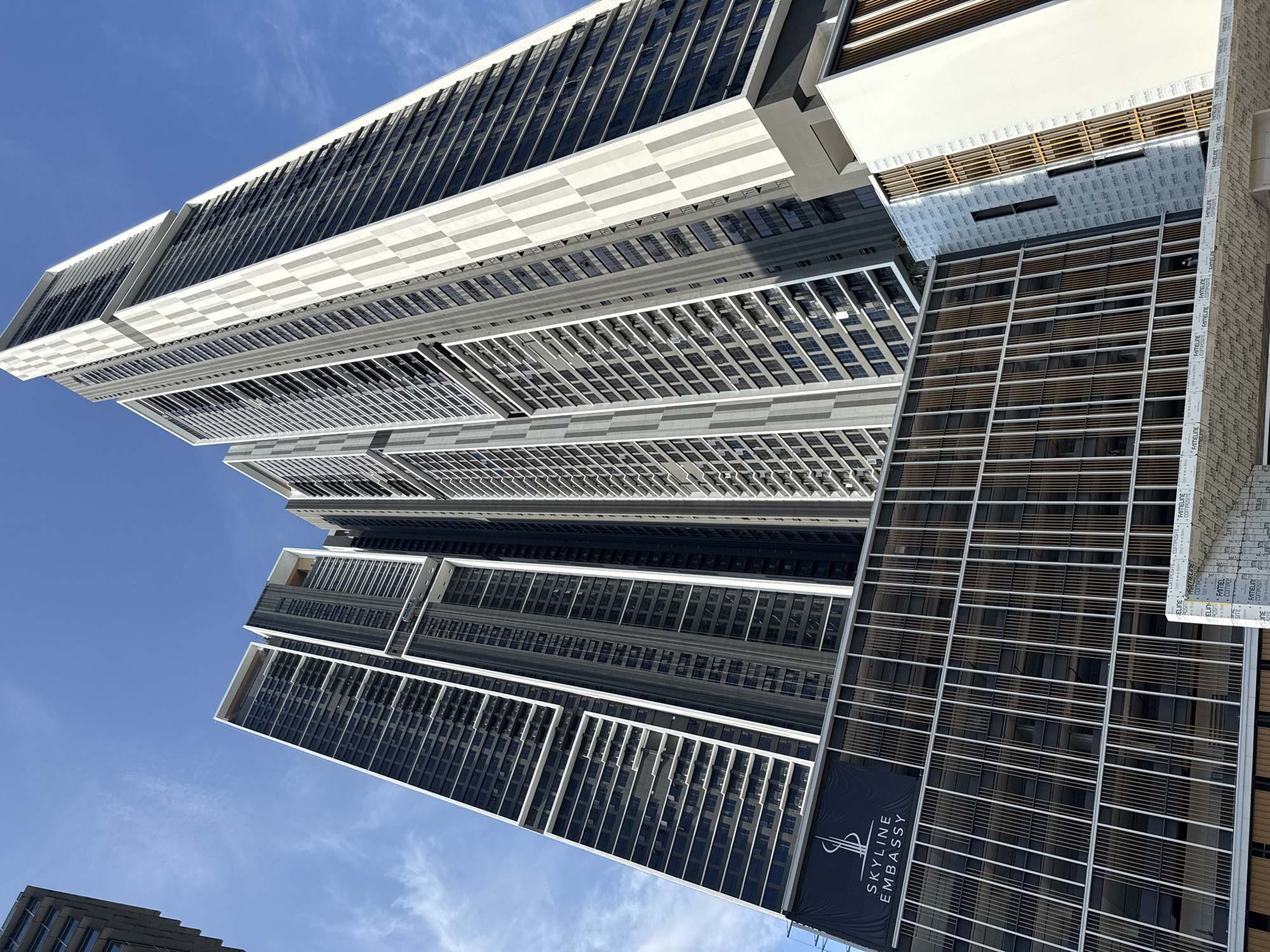 The image shows a view looking up at the Skyline Embassy building in Kuala Lumpur, Malaysia. The building is a modern high-rise with a distinctive stepped design. The facade is made up of glass and metal panels, with horizontal slats that appear to provide sun shading. A banner with the text "SKYLINE EMBASSY" is visible on one section of the building. The sky is blue with some wispy clouds. Construction materials such as cladding are also visible at the side of the building, potentially indicating an unfinished portion.