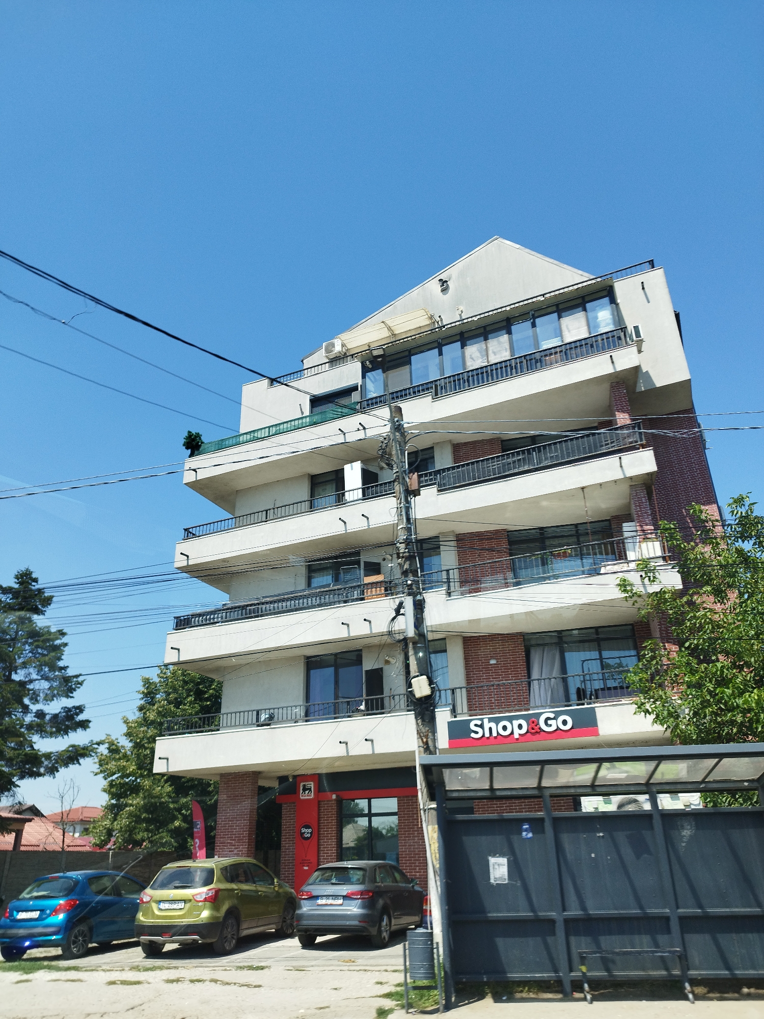 A low-angle, wide shot captures a modern, multi-story building in Bragadiru, Romania, under a clear blue sky. The building features a mix of white and exposed brick facade, with multiple balconies and large windows. A prominent "Shop & Go" sign is visible on the lower level, indicating a retail establishment. In front of the building, a bus stop shelter with dark metal framing and a clear roof stands. Three cars are parked in front of the building: a blue hatchback on the left, a lime green SUV in the middle, and a gray sedan on the right. Overhead power lines crisscross the frame, and green trees and foliage surround the building, suggesting an urban or suburban setting. The ground in front of the cars appears to be a light-colored gravel or asphalt surface.