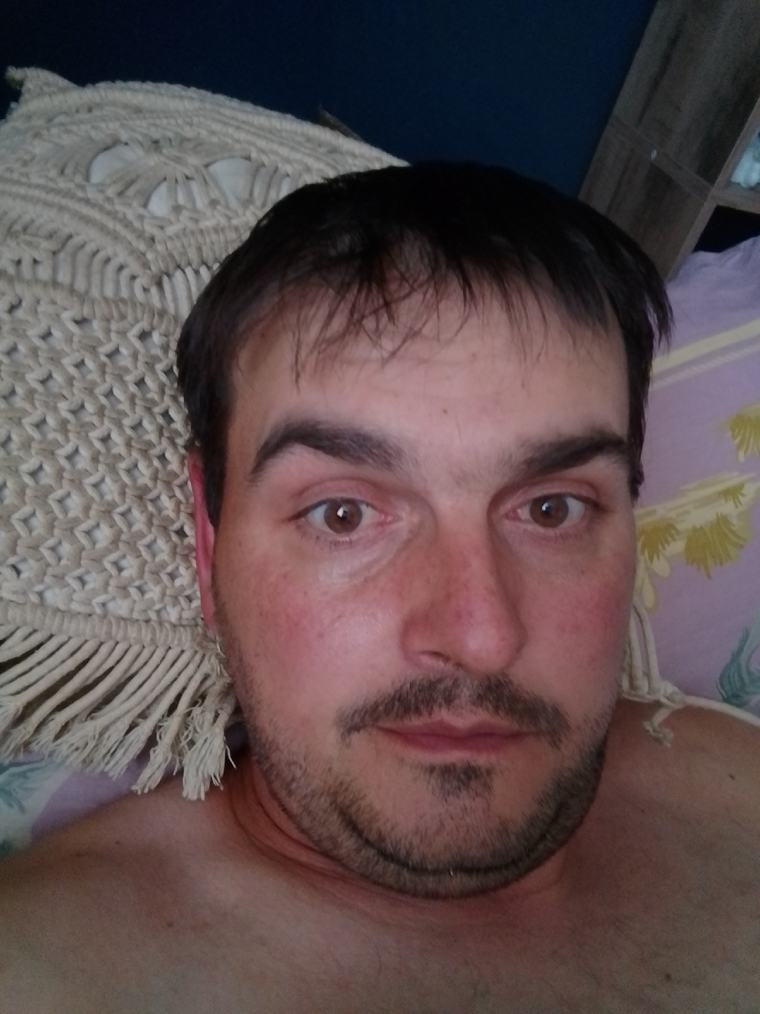 A man is lying down, looking at the camera. He has dark hair, brown eyes, and a mustache and beard. He is wearing a macrame headboard. The background is a purple and yellow floral patterned sheet. There is a wooden shelf in the upper right corner. The man is shirtless.