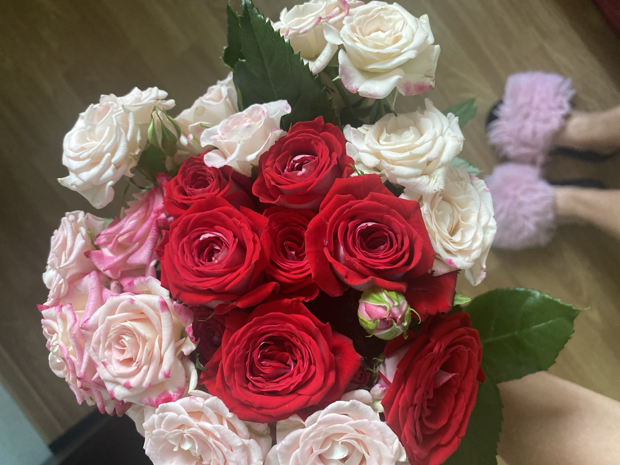 A bouquet of red and pink roses is held by a person's hand. The roses vary in bloom and color, with some fully open and others partially closed. The background appears to be a wooden floor and a wall, suggesting an indoor setting. In the lower right corner of the image, a pair of pink fluffy slippers are visible, indicating the presence of at least one person. The scene is well-lit, with natural light casting shadows. There is no indication of the city of Sochi, Russia.