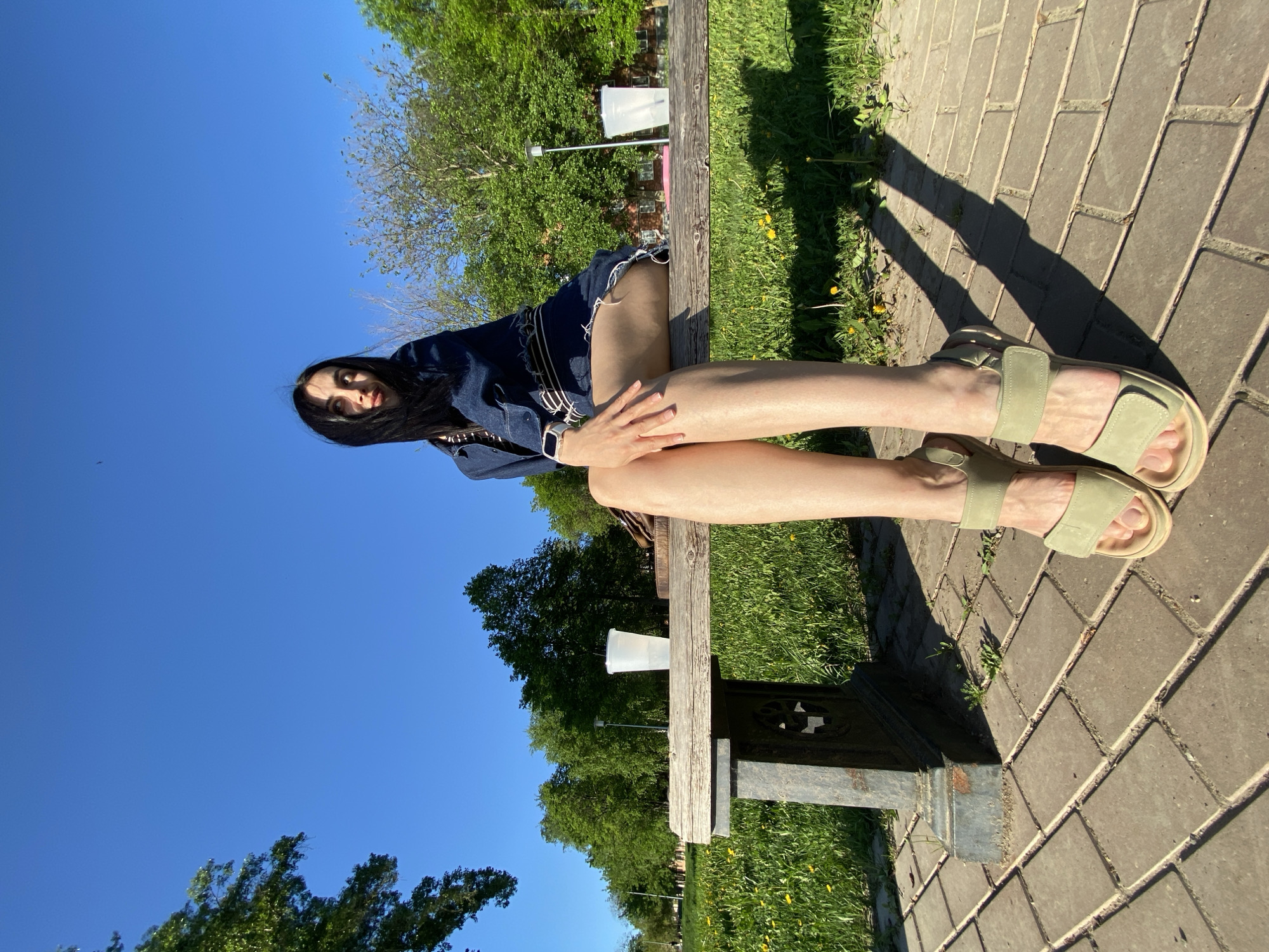 A woman sits on a wooden bench in a public park in Votkinsk, Russia. She wears a dark blue denim jacket and matching denim shorts. Her legs are crossed and she is wearing pale green sandals. The bench sits on a brick sidewalk and green grass. A clear blue sky and green trees are visible in the background. A white disposable cup sits on the bench next to the woman.