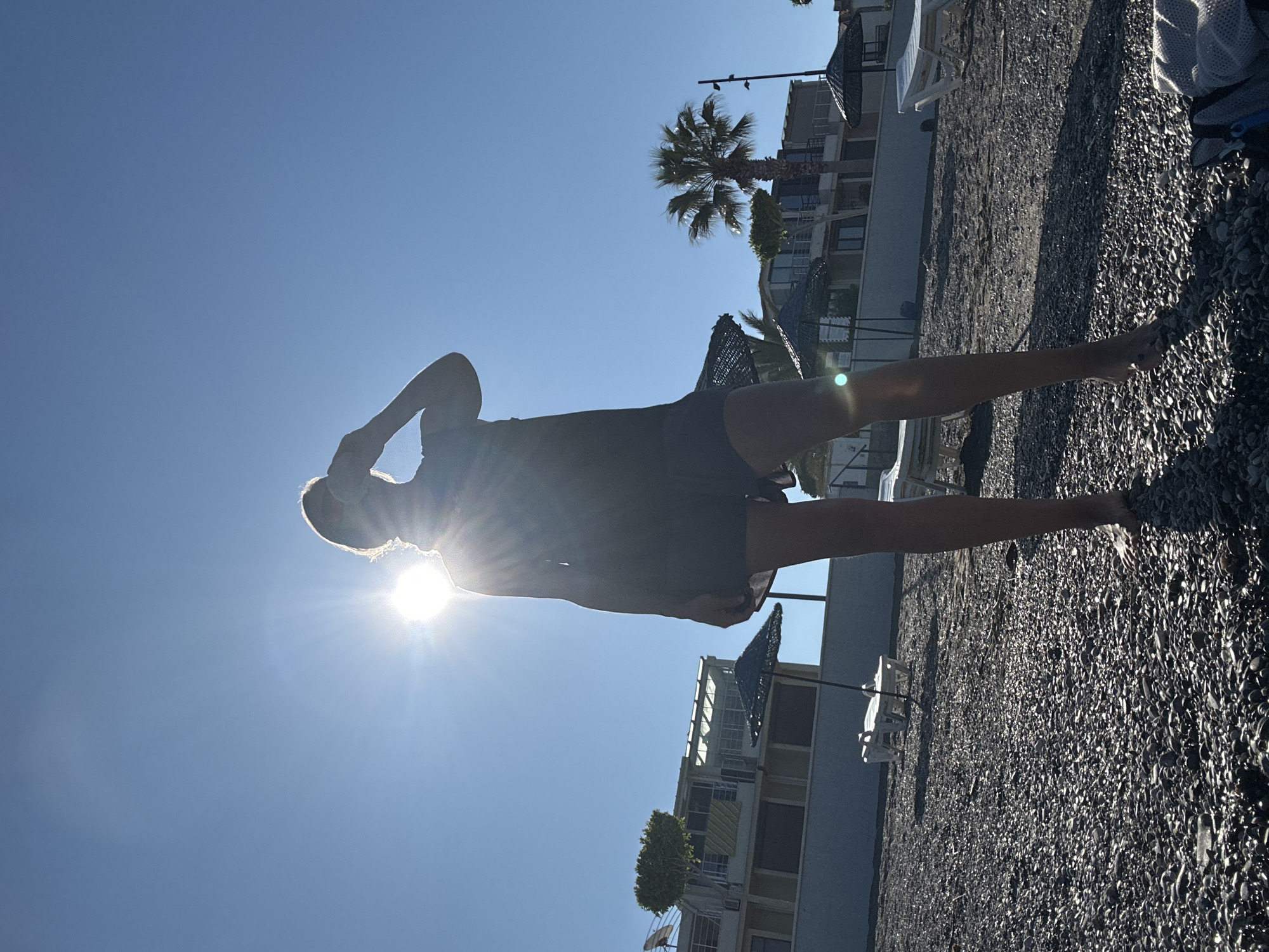 The image shows a person standing on a pebbled beach in Fethiye, Turkey, with a sunny blue sky above. The person is silhouetted against the sun, with one hand raised to their head. In the background are buildings, a palm tree, beach umbrellas, and lounge chairs. The beach pebbles are visible in detail.
