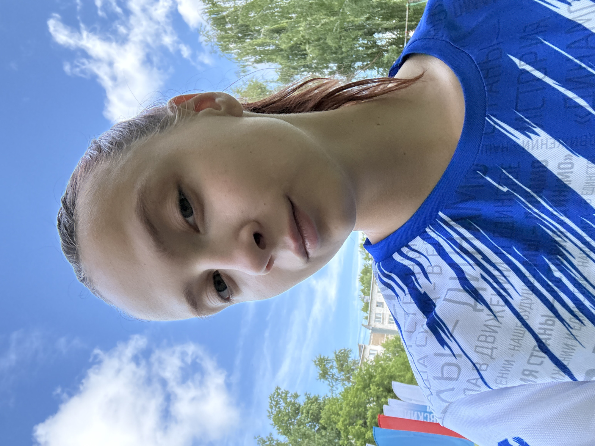 A young woman with light brown hair pulled back in a ponytail is taking a selfie outdoors against a bright blue sky with scattered white clouds. She is wearing a blue and white athletic shirt with a graphic pattern. In the background, there are green trees, some buildings, and several flags that appear to be red, white, and blue. The setting is consistent with an outdoor event or public space in the city of Chaikovsky, Russia.
