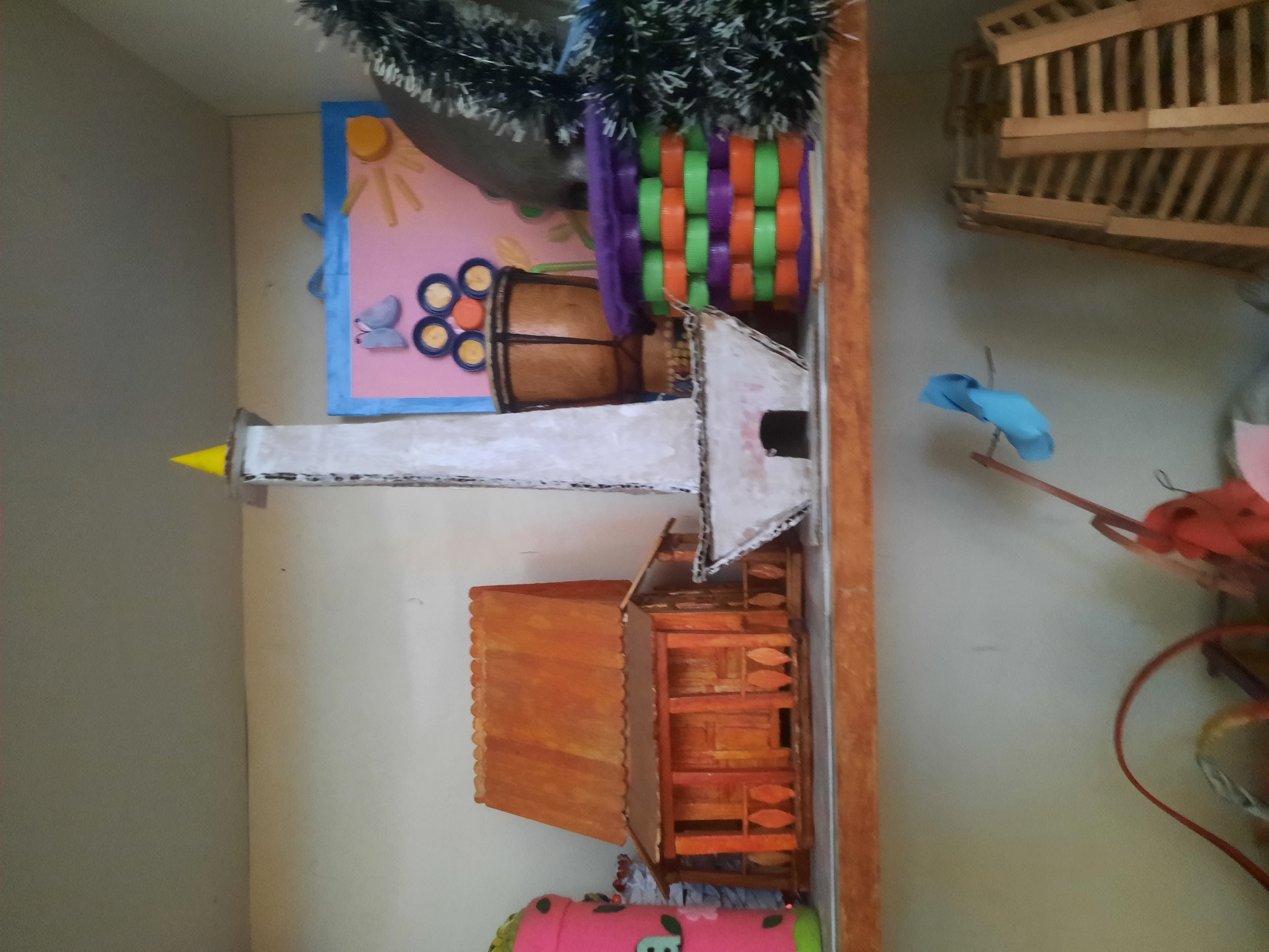 The image shows a shelf filled with various craft items. On the top shelf, there are several handmade objects. On the left is a pink cylinder with green and black decorations, followed by a miniature wooden house made of popsicle sticks. In the center is a tall, white, tower-like structure, possibly a model of a monument, topped with a yellow cone. Next to it is a small drum and a colorful craft with purple, green, and orange layers, resembling a woven basket made of bottle caps. Behind these items, a pink board with a blue border is visible, decorated with a sun and butterflies made from craft materials. A dark, fuzzy object, possibly a Christmas decoration, is on the far right. The bottom shelf contains several items, including a red wire, a blue paper airplane, and a wooden structure that looks like a folded ladder or screen. The setting appears to be a room or storage area with white walls and wooden shelves. The specific location in North Jakarta, Indonesia, cannot be determined from the image alone.