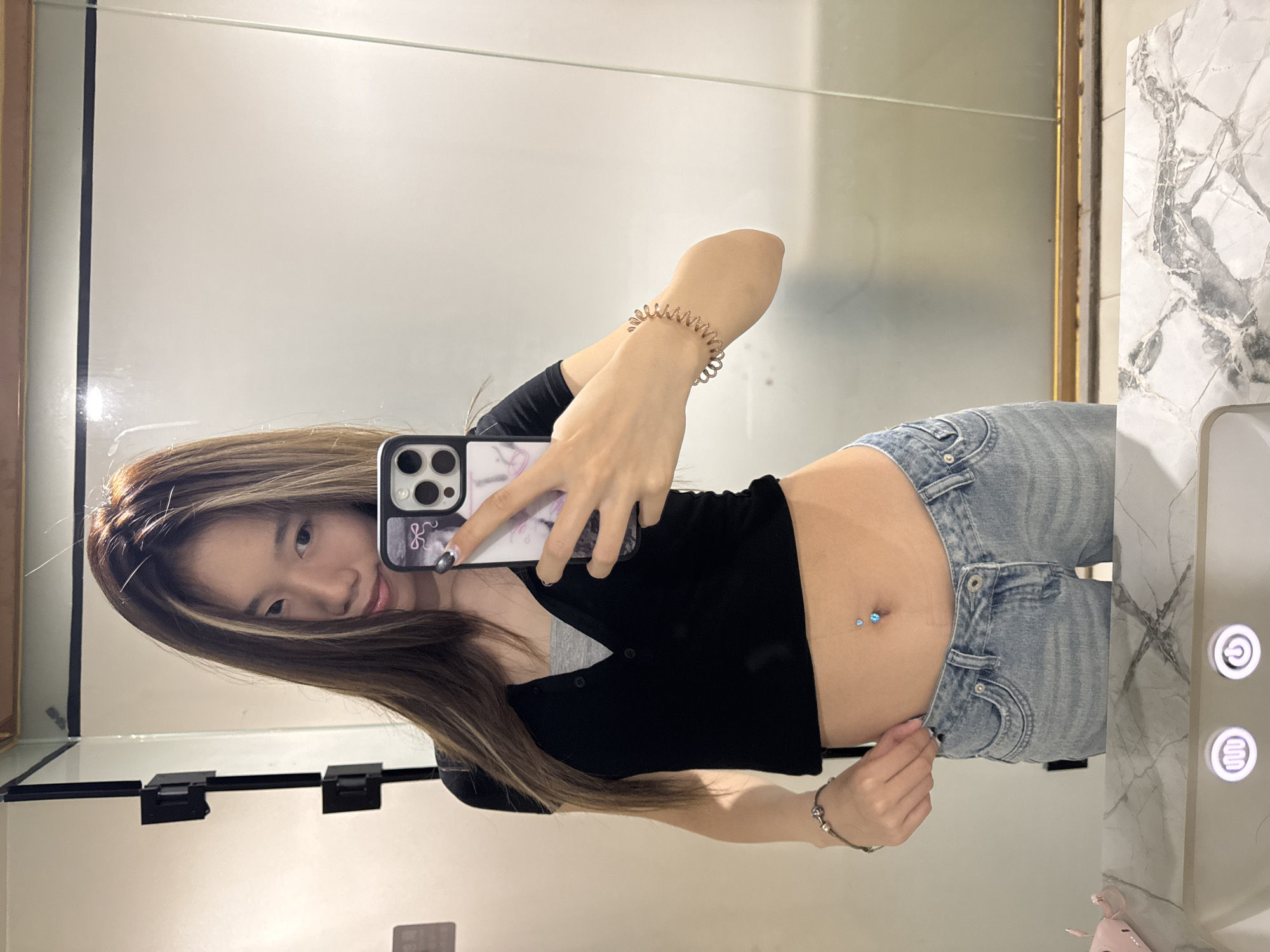A young woman is taking a mirror selfie. She is standing in what appears to be a bathroom. She is wearing a black cropped cardigan, light-wash denim jeans, and a belly button ring. She is holding a smartphone in her left hand, covering part of her face. The environment appears to be a modern bathroom with a white countertop and a mirror with a gold frame. There is no information to confirm that it is Yuzhong District, China.