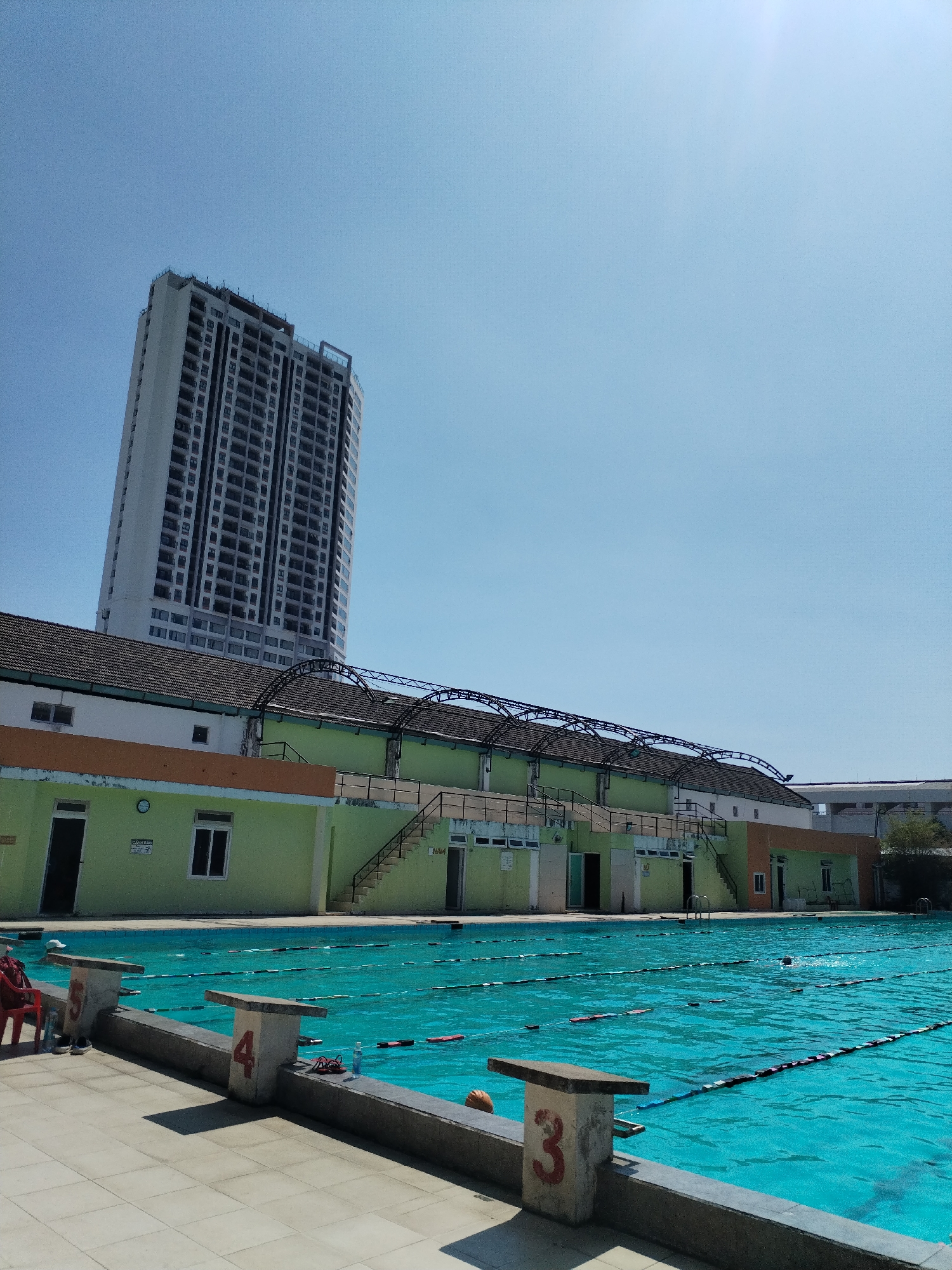 The image depicts an outdoor swimming pool located in Nha Trang Ward, Vietnam, under a clear blue sky. The pool water is a vibrant turquoise color, with black lane dividers visible. A swimmer is present in one of the lanes. The pool is bordered by a concrete deck with starting blocks numbered '3' and '4' visible.
Behind the pool is a low, single-story building painted in shades of light green and cream, featuring windows, doors, and stairs leading to the roof. A metal structure arches over part of the building's roof. In the background, a tall, modern apartment building rises into the sky. A red plastic chair is present on the pool deck.