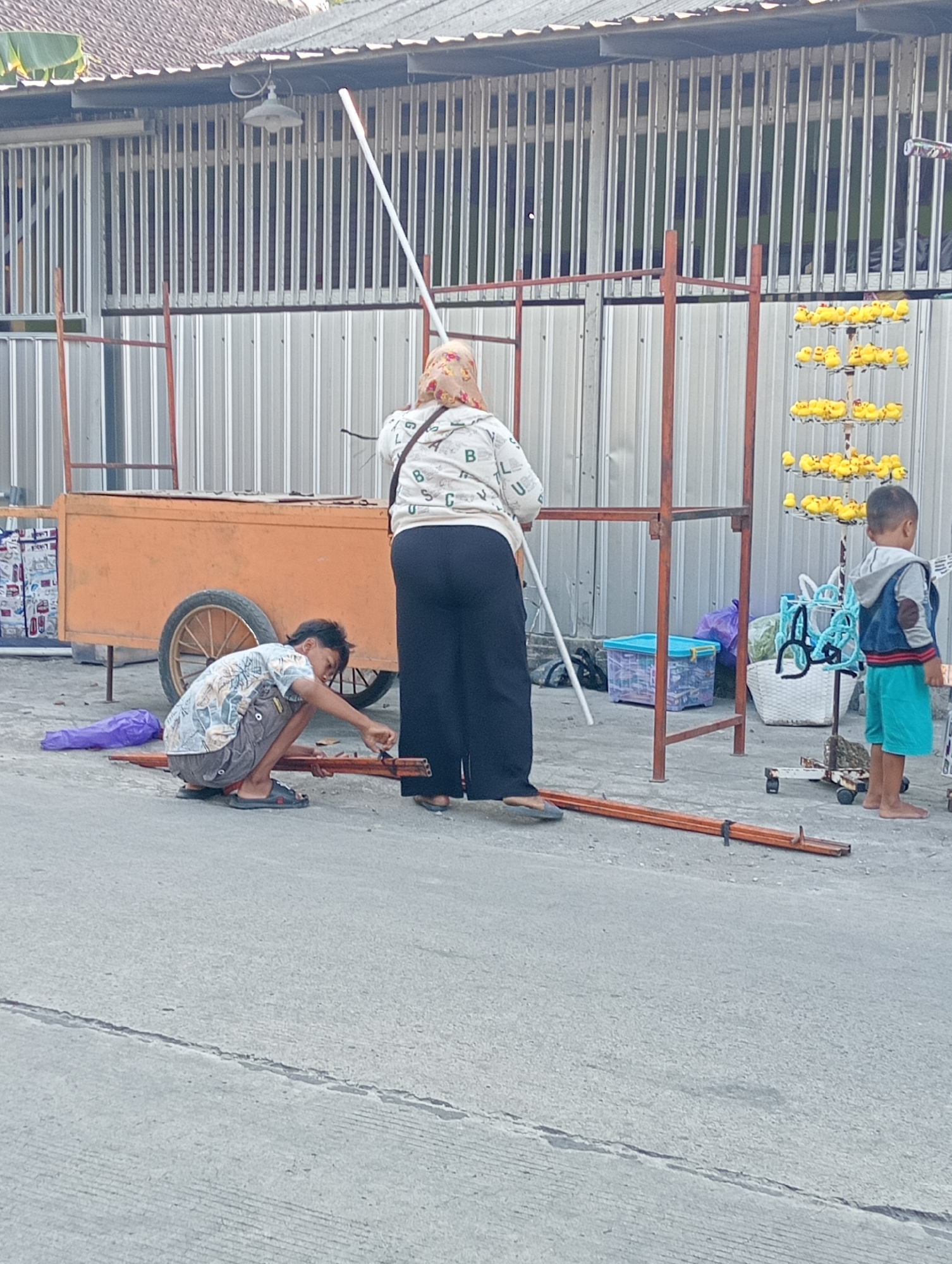 In Tanjunganyar, Indonesia, a young boy kneels on a concrete surface near an orange cart, appearing to work on disassembled wooden poles. He wears a patterned short-sleeved shirt and dark shorts. A woman in a headscarf and a white shirt with black lettering stands behind him, facing away from the camera and holding a white pole. To the right, a young boy in a blue hoodie and teal shorts stands barefoot. The background features a corrugated metal wall with vertical slats and an awning. Several metal frames are present, along with stacks of yellow rubber ducks on a rack, plastic containers, and a woven basket.