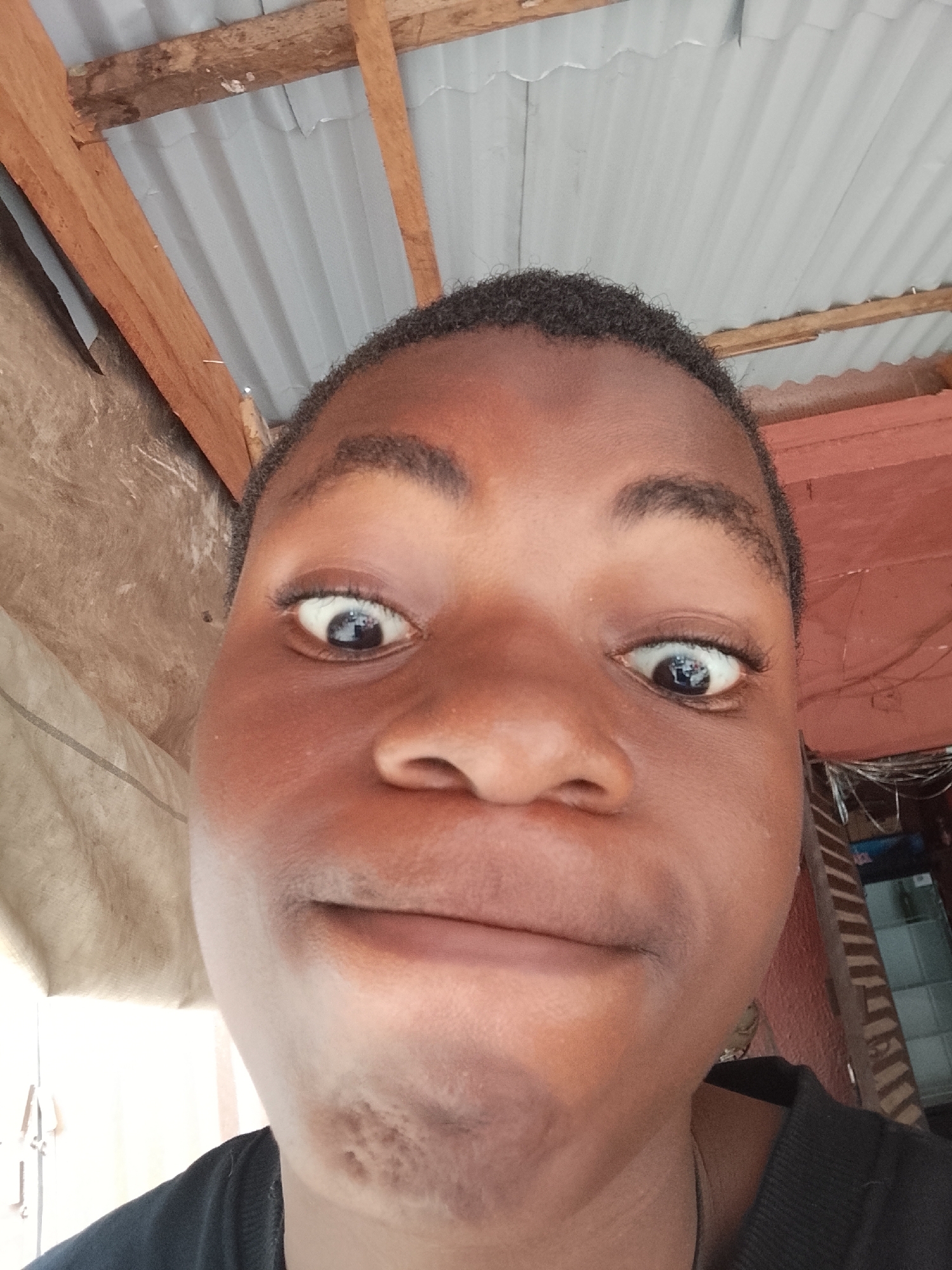 A selfie of a person with dark skin and short, dark hair is positioned low, looking upwards. The person's eyes are wide, and their mouth is slightly pursed in a humorous expression. The background consists of corrugated metal roofing supported by wooden beams. To the left, a wall with a weathered, possibly painted surface is visible, while to the right, a glimpse of what appears to be a building interior or adjacent structure can be seen. Location: Unknown, Unknown.