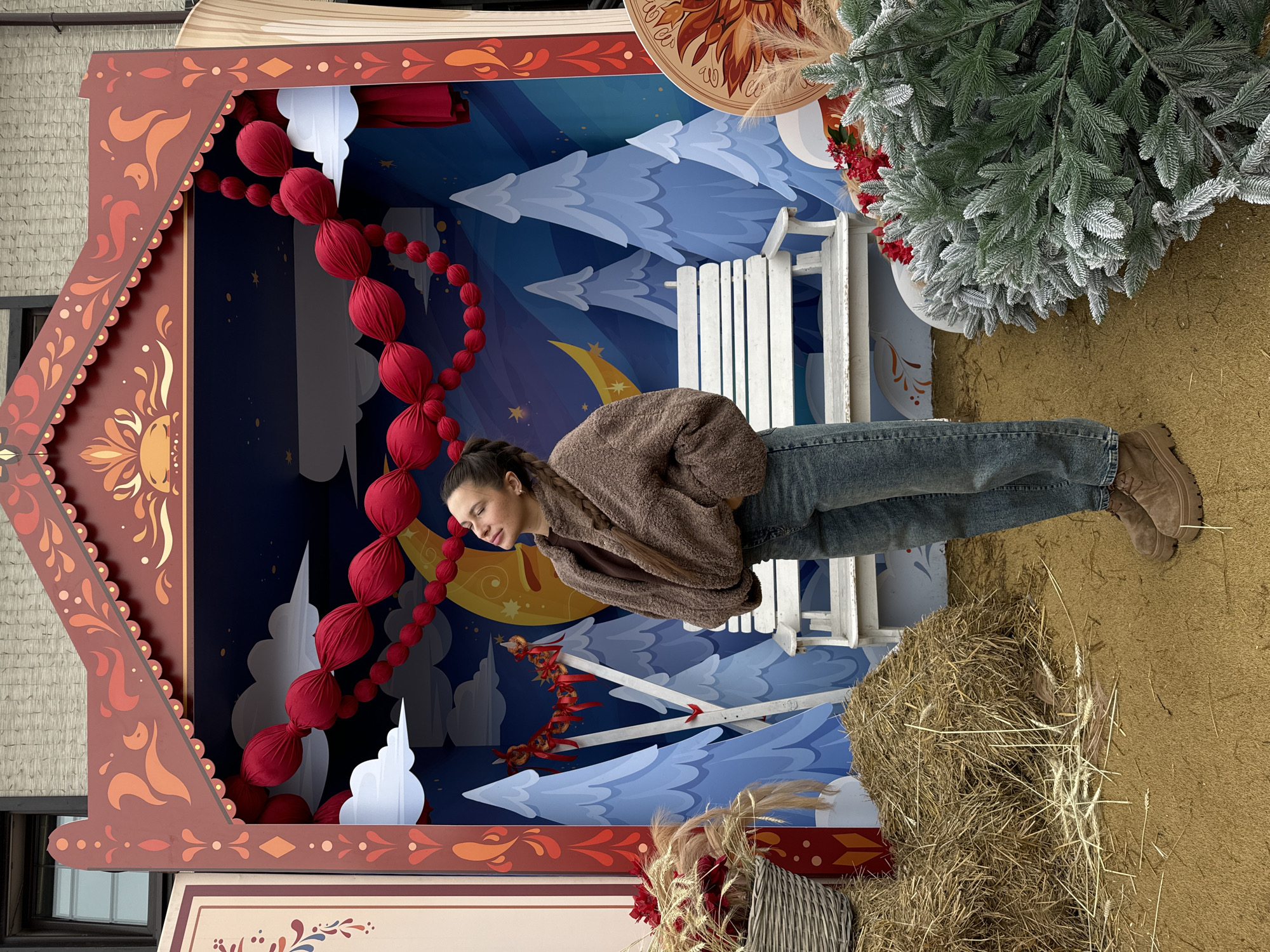 A female subject stands centered within an outdoor decorative photo display in Odintsovo, Russia. The display features a dark red wooden facade, ornately carved with traditional patterns, resembling a small house with a scalloped roofline. The interior backdrop is a painted scene depicting a stylized winter night with a large yellow crescent moon, stars, and multiple blue fir trees with white snow caps. A garland of large red fabric spheres is strung across the top of the interior, while a smaller red garland with ribbons hangs on the left side of the backdrop. A white wooden slatted bench is positioned centrally within the display.
The female, of medium build with dark hair styled in a braid over her left shoulder, wears a brown fuzzy jacket over a darker brown top, blue denim jeans, and brown platform boots. She stands with her eyes closed and head tilted slightly upward and to the left, her hands in or near her pockets. The ground of the display is covered with a tan, textured material, interspersed with straw and dried wheat stalks. To the left foreground, a light-colored woven basket filled with dried wheat and red flowers rests on a pile of hay. To the right foreground, a pile of artificial, snow-flocked green pine branches and additional red decorative elements are arranged, partially obscuring a circular decorative object with an abstract pattern.
