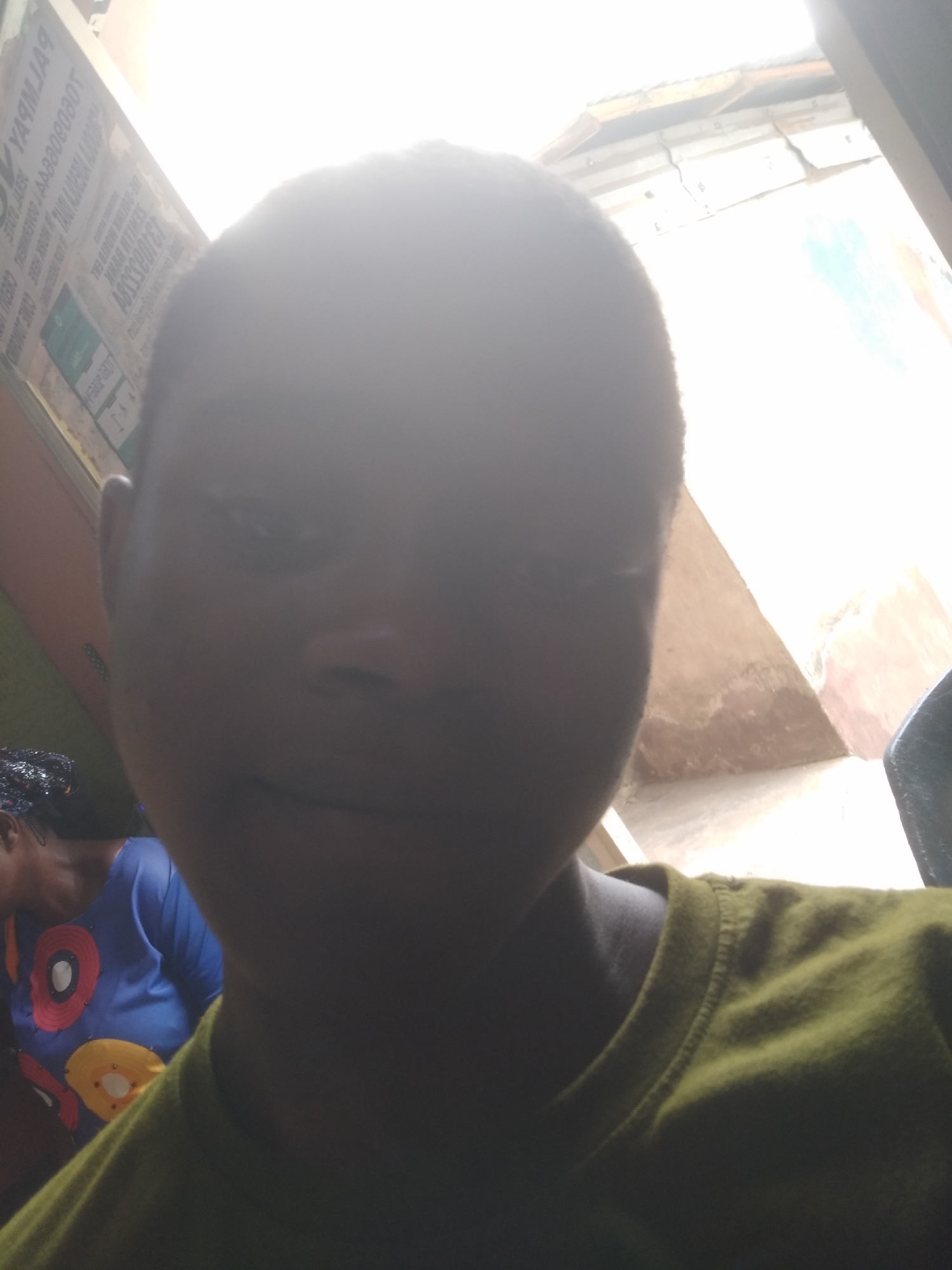 A close-up selfie of a person with short hair, wearing an olive-green t-shirt. The background is overexposed, showing a structure with what appears to be corrugated metal roofing. A woman wearing a blue shirt with circular patterns is partially visible in the lower left corner. A sign with text in a foreign language is seen to the left of the person. The location is likely Shangisha, Nigeria, based on external information.