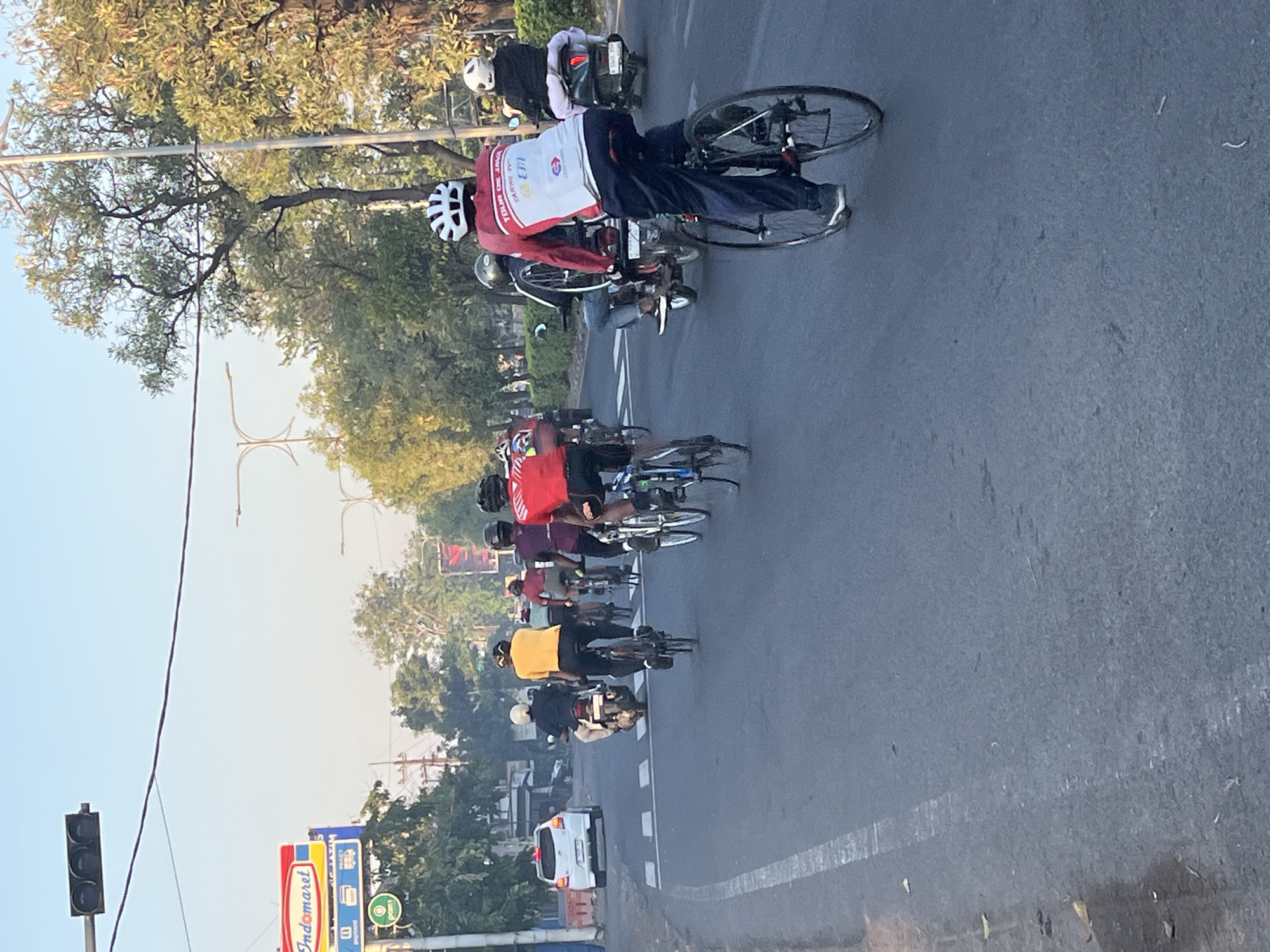 The image shows a road with several cyclists and a few motorcycles. In the foreground, a person in a red and white cycling jersey and dark pants is riding a bicycle. Behind them, another cyclist in a purple jersey is visible. Further ahead, there are multiple cyclists wearing helmets and athletic attire, some in red and yellow jerseys. Two motorcycles are also present on the road, one carrying two individuals and another carrying a single person. On the left side of the road, a white car is parked or stopped. A traffic light is visible overhead, and a prominent "Indomaret" sign is present on the left, indicating a convenience store. Trees line the side of the road, and the sky is clear and bright, suggesting it is daytime. The scene appears to be a street in Kludan, Indonesia, likely during a cycling event or group ride, as indicated by the coordinated cycling attire of several individuals.
