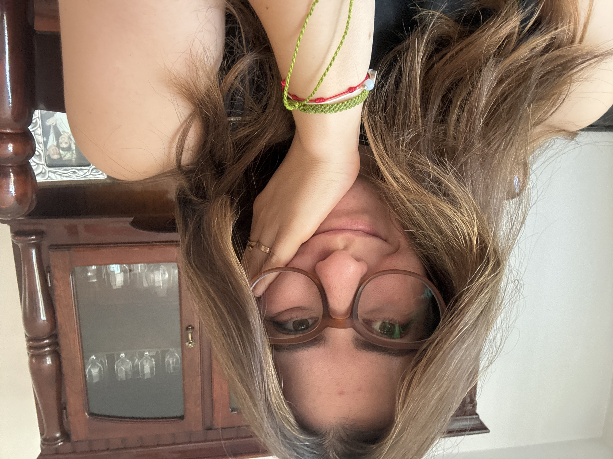 A woman with light brown hair, wearing glasses, is looking at the camera. She is resting her head on her hand, which has a gold ring. She is wearing bracelets on her wrist. Behind her is a wooden cabinet with glassware visible inside. The location is likely Puebla, Mexico, due to the type of furniture.