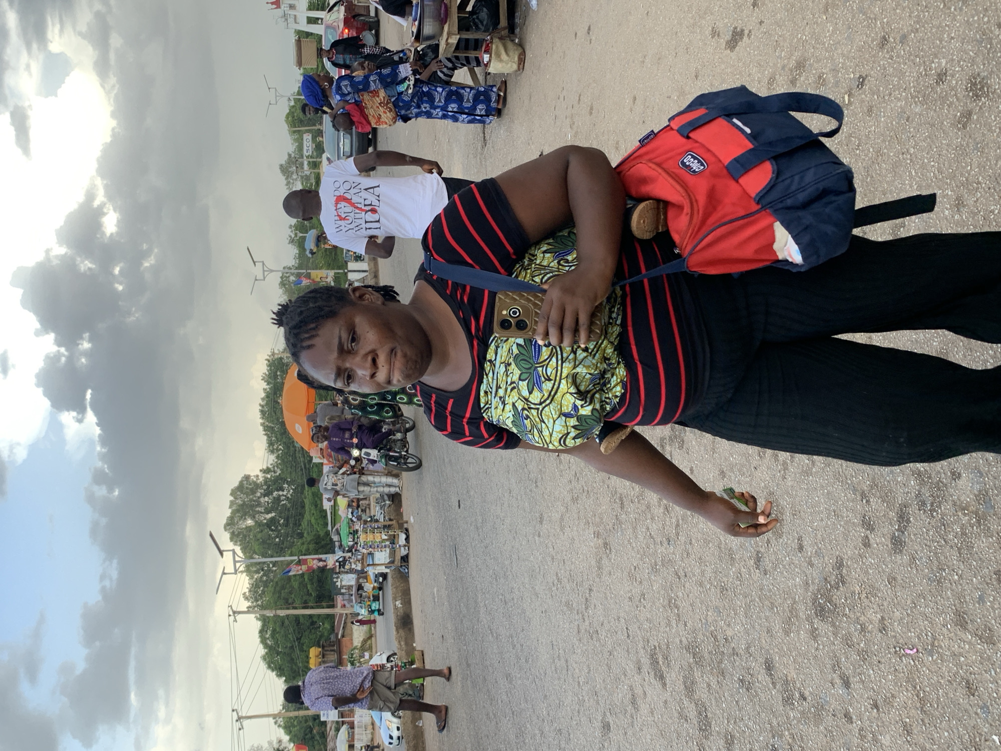 A woman with dark skin and braided hair walks in the foreground, carrying a red and blue bag over her left shoulder and holding a smartphone in her right hand. She is wearing a black and red striped t-shirt over a colorful patterned fabric, and black pants. A small child is strapped to her chest with the patterned fabric. The background shows a street scene with various people, motorcycles, and market stalls. A man in a white t-shirt with text on the back is walking away from the camera in the middle ground. Further in the background, there are buildings, trees, and a cloudy sky. The ground is paved with asphalt. The scene appears to be a bustling street market in Jos, Nigeria.
