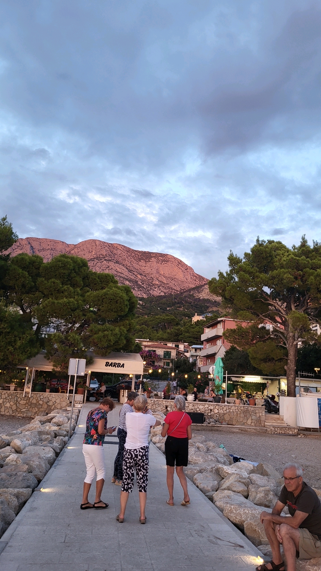 In Općina Tučepi, Croatia, at dusk, a group of four individuals, three women and one man, are visible on a paved walkway. The three women are standing and facing away from the camera, with one woman on the left wearing a floral top and white pants, another in the center wearing a striped shirt and patterned capri pants, and a third on the right in a red top and black shorts. The man is seated on large rocks to the right of the walkway, wearing a dark t-shirt and khaki shorts.
Behind the walkway, a beach with scattered rocks leads to a seaside promenade lined with trees and buildings. A restaurant with white awnings and the sign "BARBA" is prominent, with outdoor seating and string lights. In the background, a densely forested mountainside is illuminated by the setting sun, casting a pinkish glow on the rugged peaks. The sky above is cloudy with a mix of grey and lighter tones, indicating the transition from day to evening. The overall scene depicts a casual outdoor atmosphere in a coastal town.