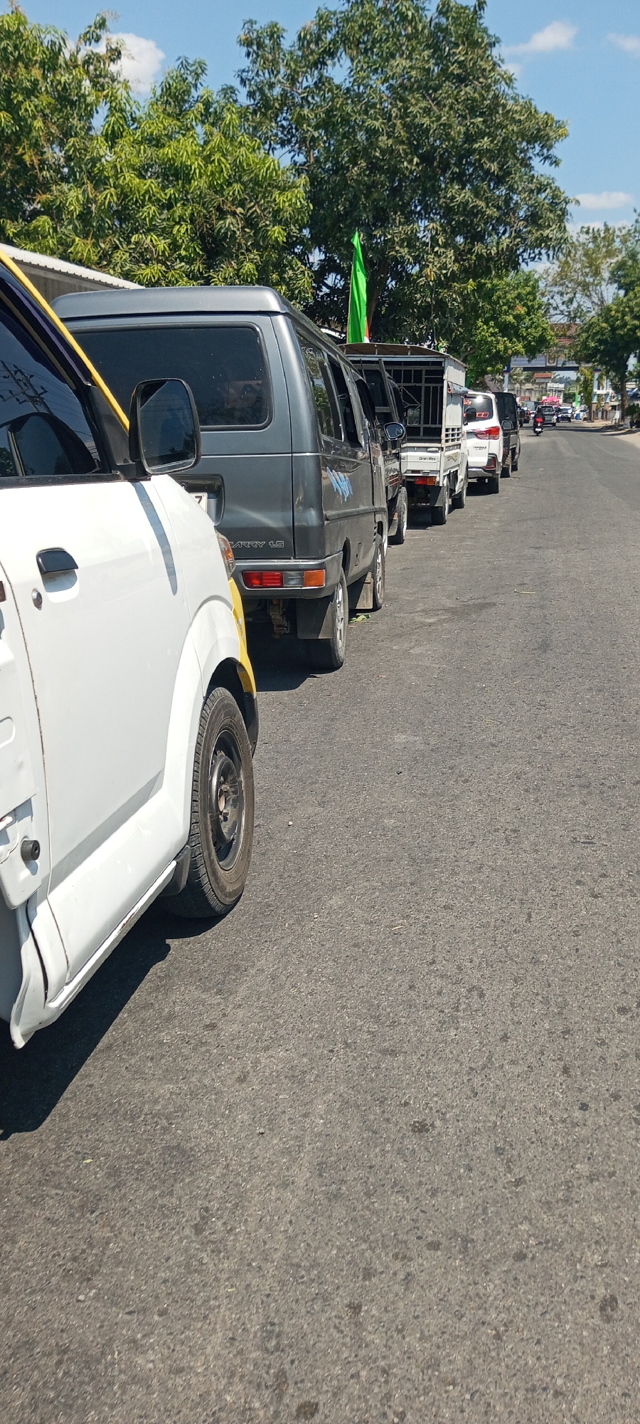 The image shows a line of utility vehicles, including vans and trucks, parked or moving slowly on an asphalt road in Lempa, Indonesia. The vehicles are positioned along the right side of the road, with one white vehicle in the foreground on the left. Trees and foliage are visible in the background, lining the road. The sky is blue with some clouds. One of the vehicles, a dark gray van, has a green flag attached to its roof. The scene suggests a calm, possibly a parade or a procession, or simply traffic on a sunny day.