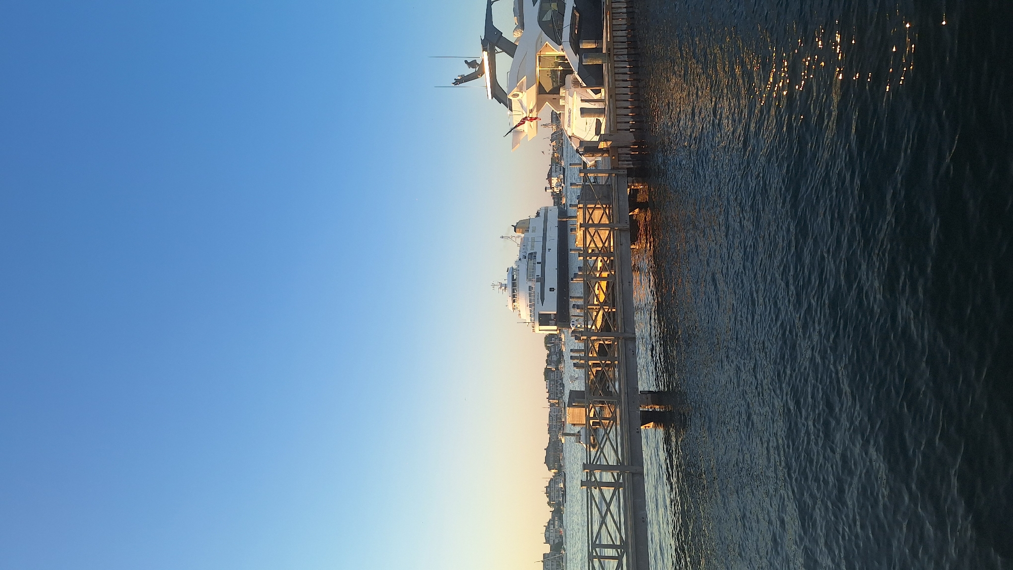 In Nantucket, Massachusetts, on a clear day with a bright blue sky, a ferry boat is docked near a pier. The ferry, white with a yellow funnel, is positioned in the middle ground. To the right, a modern white yacht with black accents is also docked. The pier extends into the water from the foreground, with a wooden railing. Buildings and trees are visible on the distant shore, suggesting a waterfront town. The water is dark blue with reflections of the setting sun.