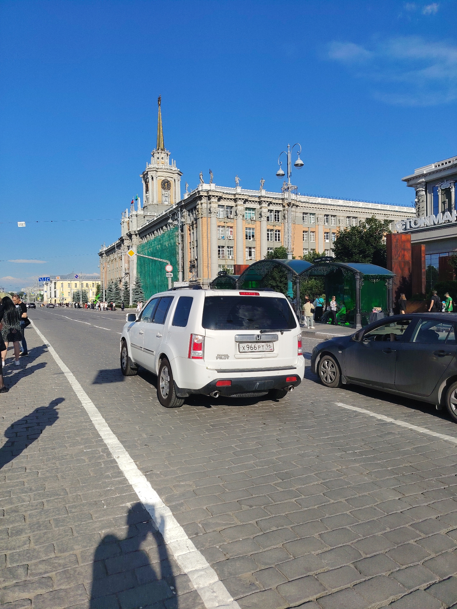 The image depicts a sunny day in a city, likely Yekaterinburg, Russia, given the location context. The scene is a street with cobblestone paving. In the foreground, a white Honda Pilot SUV is parked. To its right, a dark gray sedan is also parked. A white line marks a parking or traffic lane.
Several people are visible in the mid-ground, some walking along the sidewalk and others near green, gazebo-like structures that might be bus stops or waiting areas. Shadows of people are cast on the ground, suggesting a midday time. The sky is a clear, vibrant blue, indicating fair weather.
Dominating the background is a large, ornate building with a prominent clock tower topped with a spire. The building has classical architectural elements, including columns and statues. Green netting covers a section of the building, suggesting ongoing construction or renovation. Another building to the right displays the word "ГЛОТОМАК" (GLOTOMAK) in large white letters. Traffic and more buildings are visible further down the street, indicating an urban environment.
The license plate of the white SUV is partially visible, displaying Russian characters and numbers: "X966PT 96". The overall atmosphere is bright and urban, with a sense of everyday city life unfolding.