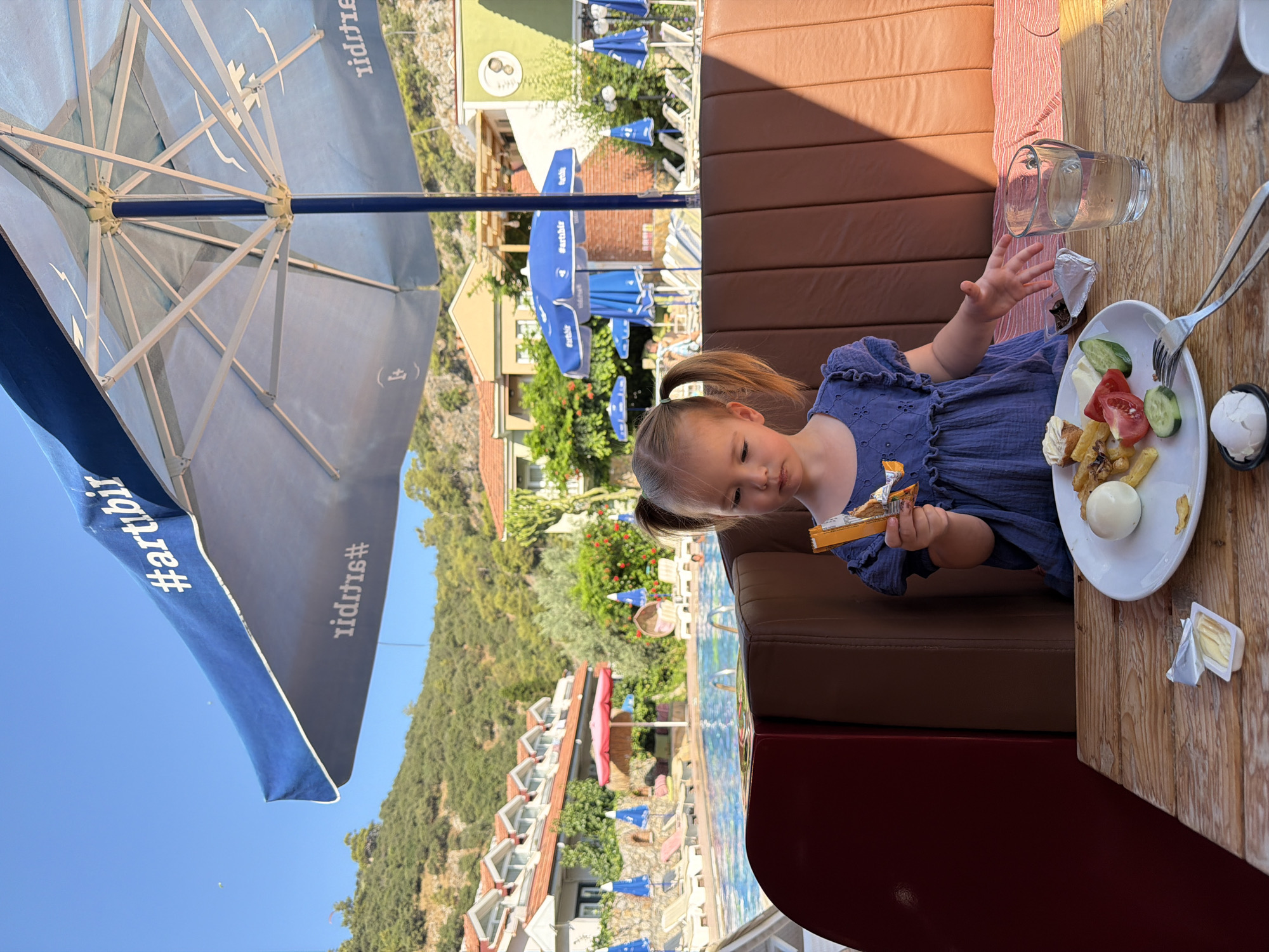 A young girl with blonde pigtails, wearing a blue dress, sits at a wooden table under a large blue umbrella with white trim. The umbrella has a white hashtag logo with the text "artibir" on it, and another similar logo mirrored on the underside. The girl holds an orange snack bar in her right hand, and her left hand is outstretched towards the table. In front of her is a white plate with a boiled egg, french fries, tomato slices, cucumber slices, and some small pastries. To the right of the plate is a clear glass of water. A small packet of butter is also on the table. The background shows a swimming pool with lounge chairs and umbrellas, and a hillside covered in lush green trees and vegetation. In the distance, there are several buildings with red-tiled roofs, typical of Mediterranean architecture. The sky is a clear, bright blue. The scene suggests a sunny day at a resort or hotel in Fethiye, Turkey.