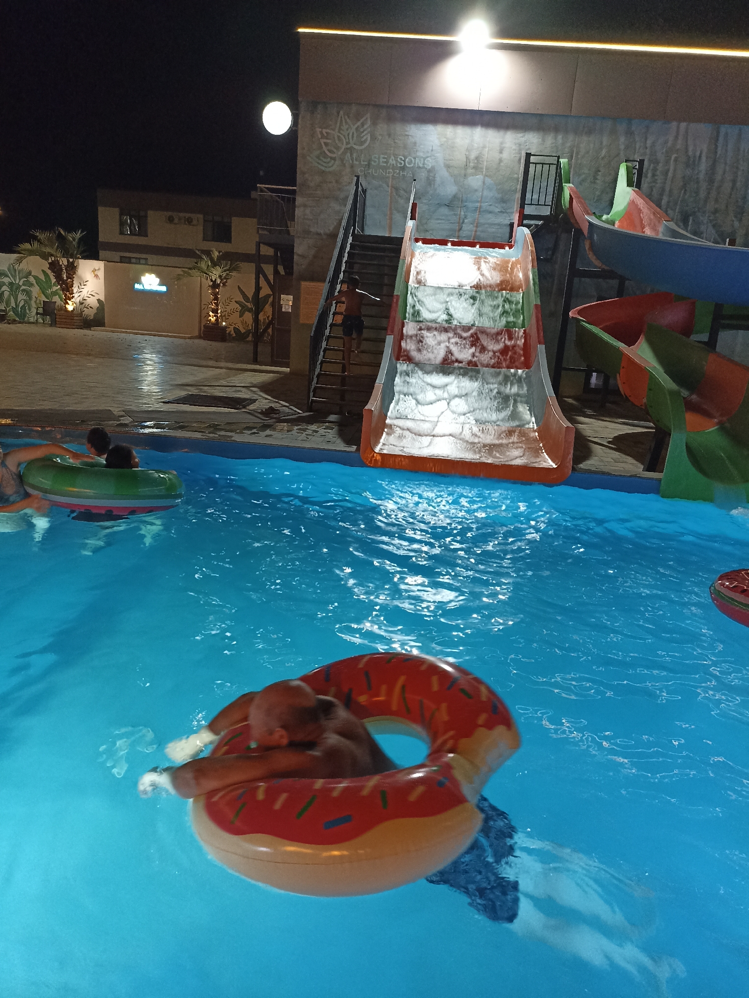At night, a person is floating on a donut-shaped inflatable in a swimming pool at the "All Seasons" resort in Sorbulak, Kazakhstan. Another person is visible in the water on a green and red inflatable ring. In the background, there is a water slide with red, green, and blue sections, and stairs leading up to it. The pool area is illuminated by artificial lights, and palm trees and a building are visible in the distance.