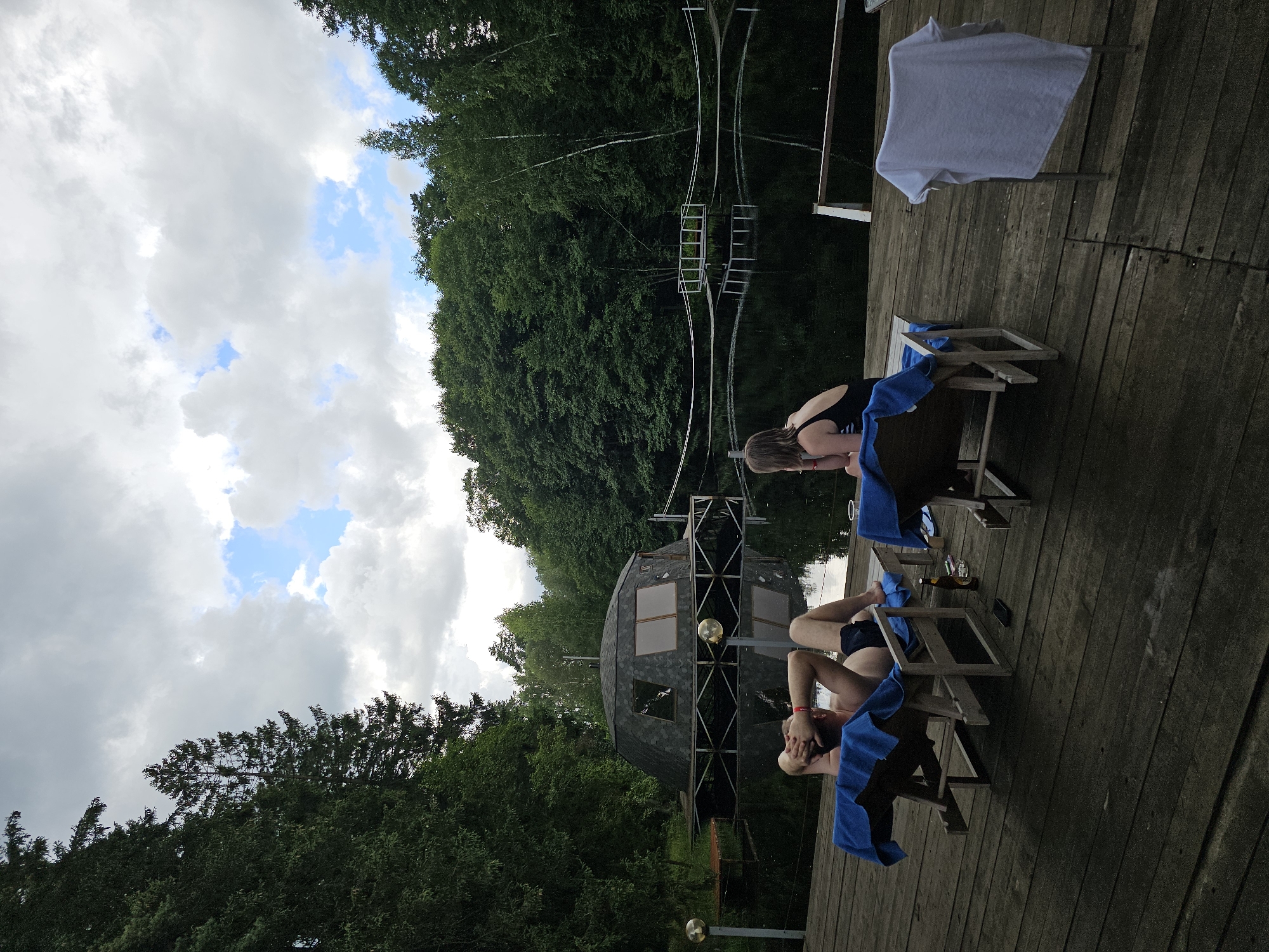 In Ulitkino, Russia, two people are relaxing on a wooden deck next to a tranquil body of water. A man reclines on a lounge chair, his hands behind his head, while a woman sits on another chair nearby, facing away from the camera. On the water, a unique, geodesic dome-shaped structure is perched on stilts. Dense, green trees surround the lake, with a cloudy sky overhead. A white t-shirt is draped over a chair on the deck, and a beer bottle and a mobile phone are placed on the wooden planks.