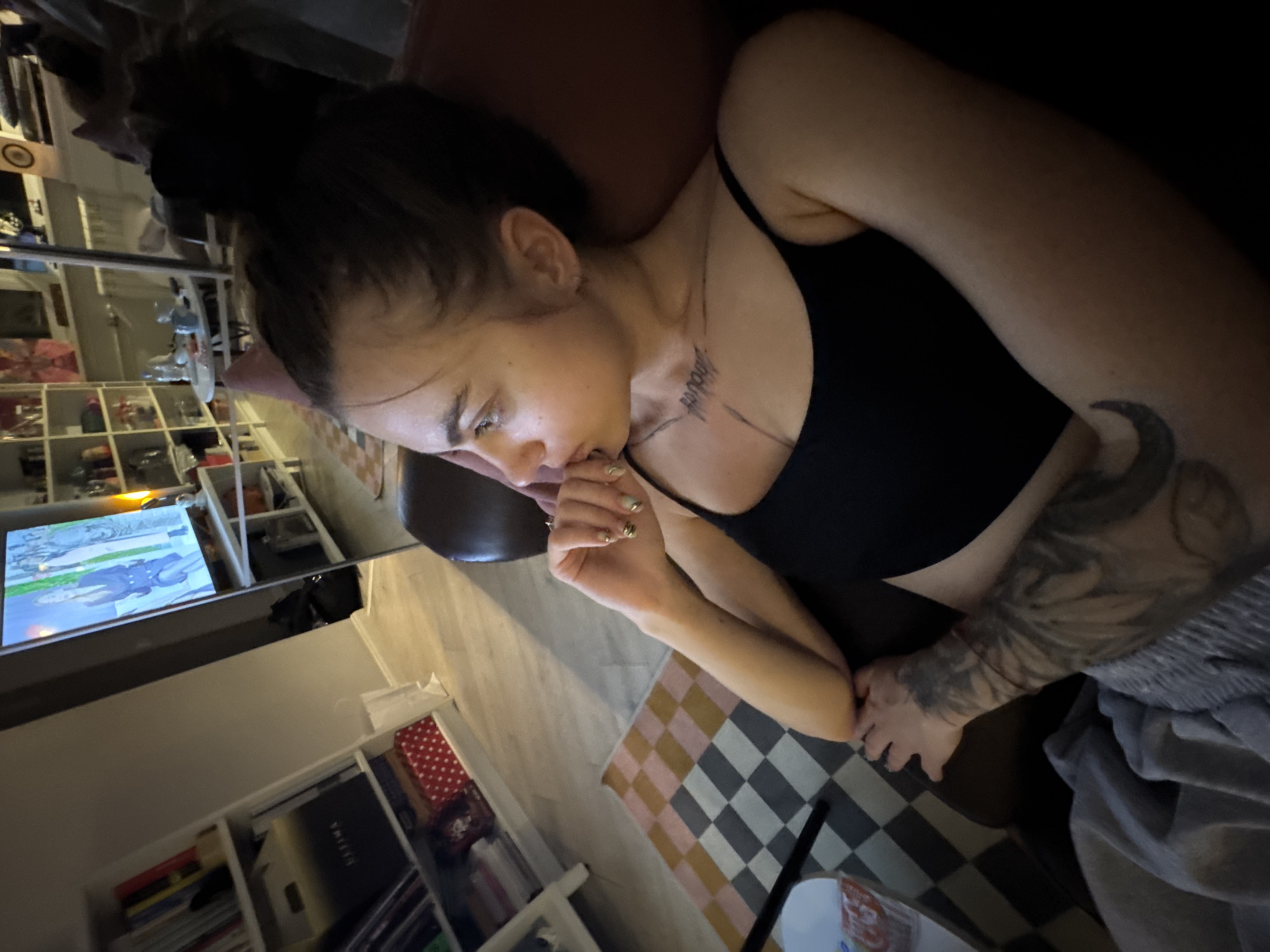 An overhead shot shows a woman from the chest up, seated on a couch, looking to her left. She has her hair pulled back in a messy bun and is wearing a black tank top. A tattoo is visible on her chest and forearm. In the background, a television screen displays an image of people, and bookshelves are filled with books and decorative items. The room appears to be a living area.