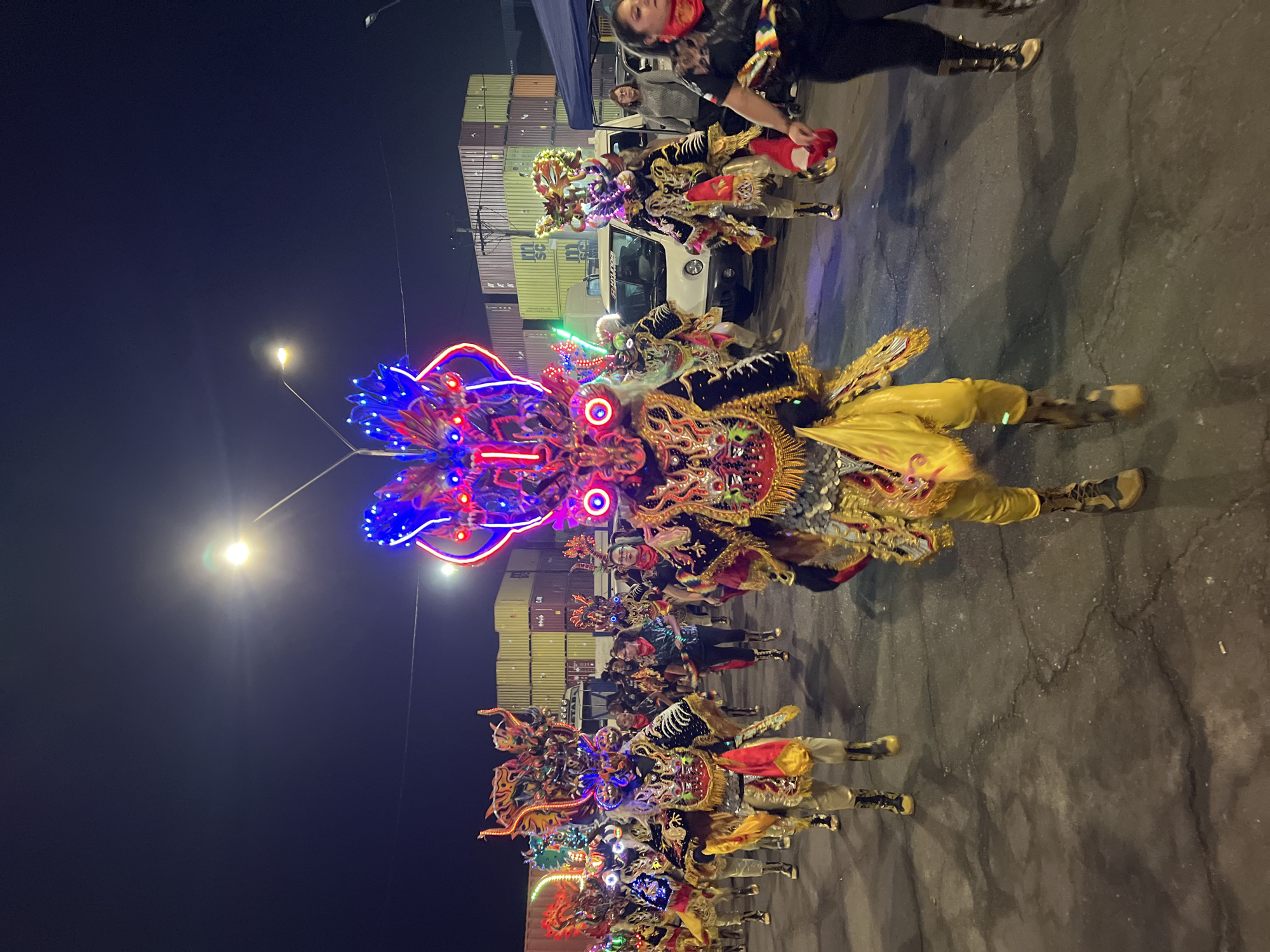 At night in Arica, Chile, a parade featuring elaborately costumed dancers proceeds along a paved surface. The dancers wear vibrant, multi-colored outfits with ornate masks, some illuminated with LED lights. Cargo containers and a white pickup truck are visible in the background. Streetlights illuminate the scene. People stand along the periphery, observing the parade. The focus is on the first row of dancers closest to the camera.