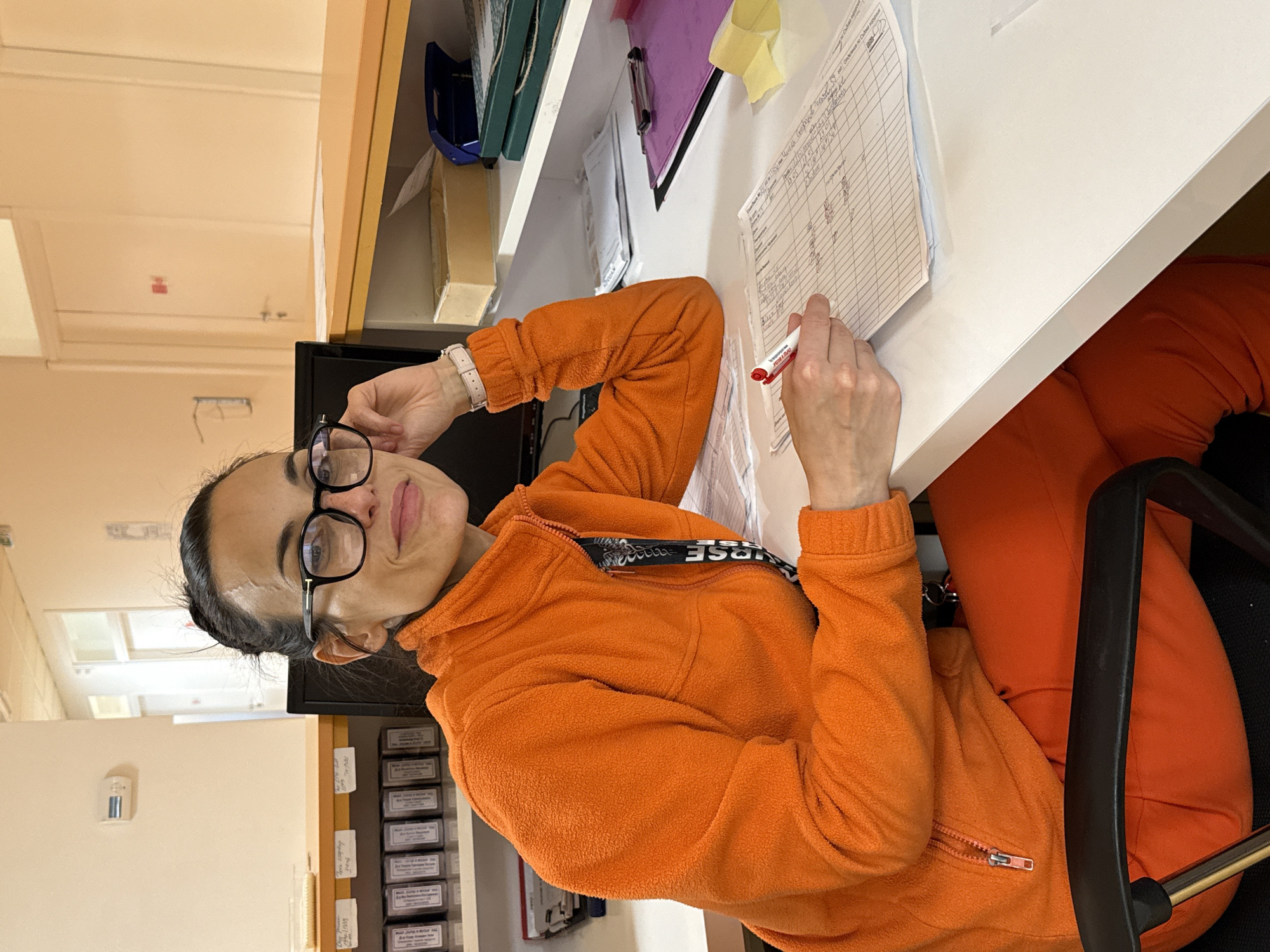 A woman is seated at a desk in an indoor setting. She is wearing glasses and an orange fleece zip-up sweater, and she is holding a red marker in her right hand, appearing to write on a document with a table format. The desk has various items on it, including papers, binders, and a computer monitor. There are drawers and labels behind the desk. The location is likely a medical office or similar setting. The city of Pleven, Bulgaria, cannot be verified from the image.