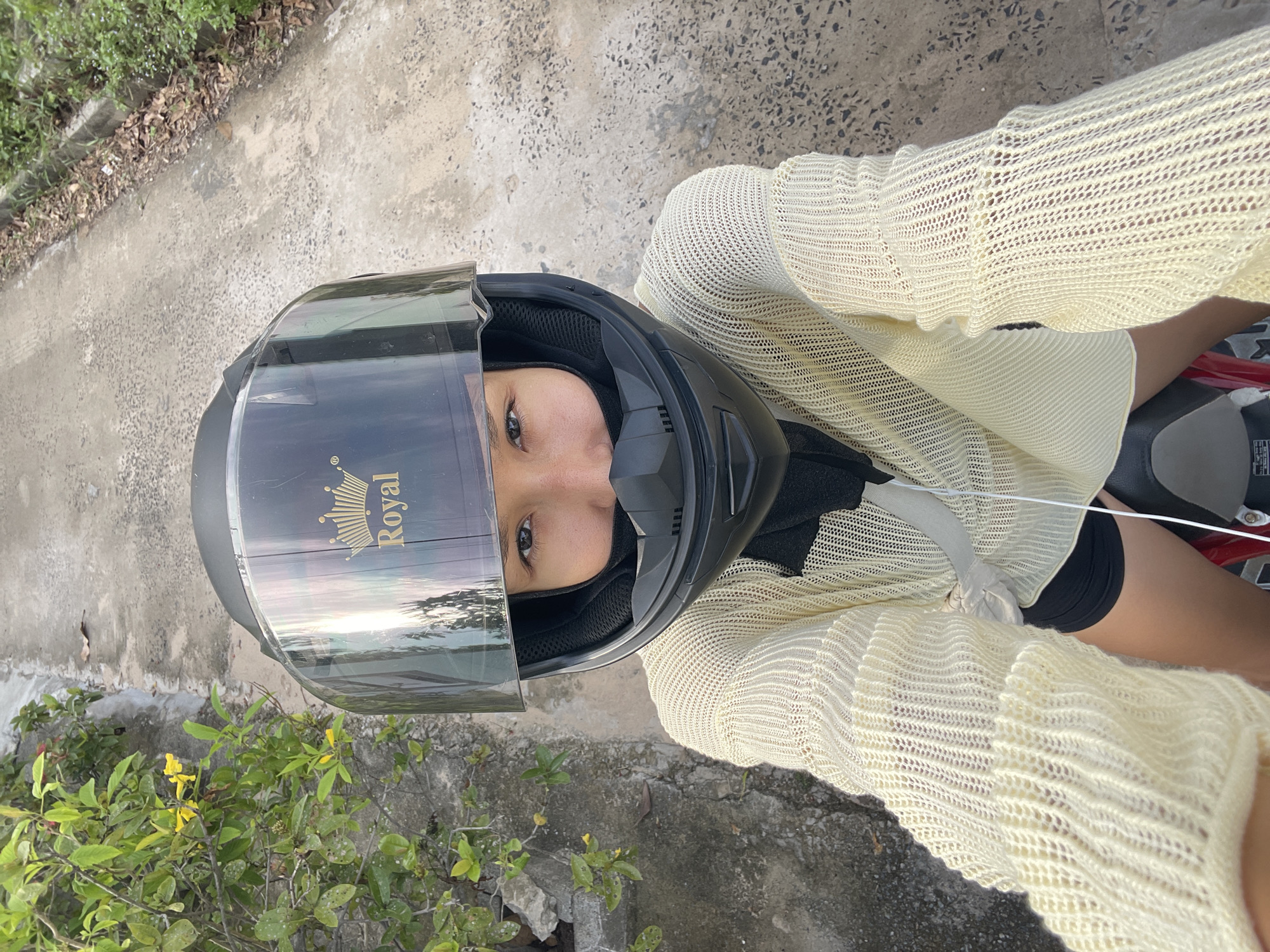 A high-angle, portrait-oriented photograph, rotated 90 degrees clockwise, captures a person wearing a full-face motorcycle helmet while sitting on a vehicle. The subject, likely female, has one eye visible through the helmet's visor, looking directly at the camera with a neutral expression.
The person is wearing a light yellow or cream-colored open-knit long-sleeve top, revealing glimpses of skin and black shorts underneath. The top has a distinct textured, mesh-like pattern. Parts of a red and grey motorcycle seat are visible beneath the person's right leg.
The black helmet features a clear visor, which reflects a bright, possibly sunset or sunrise sky with trees, suggesting it is daytime. A gold "Royal" logo with a crown icon is prominent on the visor.
The background consists of a textured concrete path or road, with green foliage, bushes, and scattered dry leaves along its edge. The overall setting appears to be outdoors in a casual, possibly roadside environment. The location context is Phu Thuy Ward, Vietnam.
