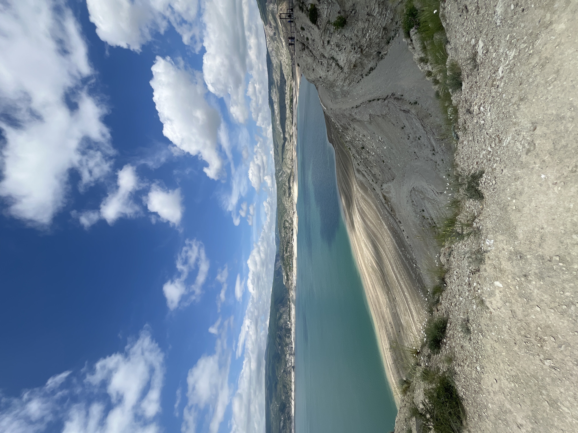 The image depicts a view of the Chirkey Dam reservoir in Chirkey, Russia, photographed from an elevated position along the embankment. The reservoir’s turquoise water stretches horizontally across the frame. To the left, the sky is visible with scattered white clouds against a blue background. On the right side of the image, a rocky embankment slopes down to the water’s edge. In the upper left, a construction or maintenance structure is visible at the edge of the frame. The image shows land erosion, and water level drop.