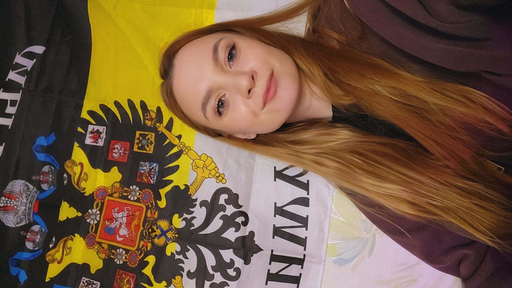 A selfie of a fair-skinned young woman with long red hair, wearing a dark purple or brown sweatshirt, posing with a flag featuring a black double-headed eagle emblazoned with a heraldic shield, and lettering. The flag has a black, yellow, and white color scheme. The background is a floral pattern. The location is Зипуново, Russia.