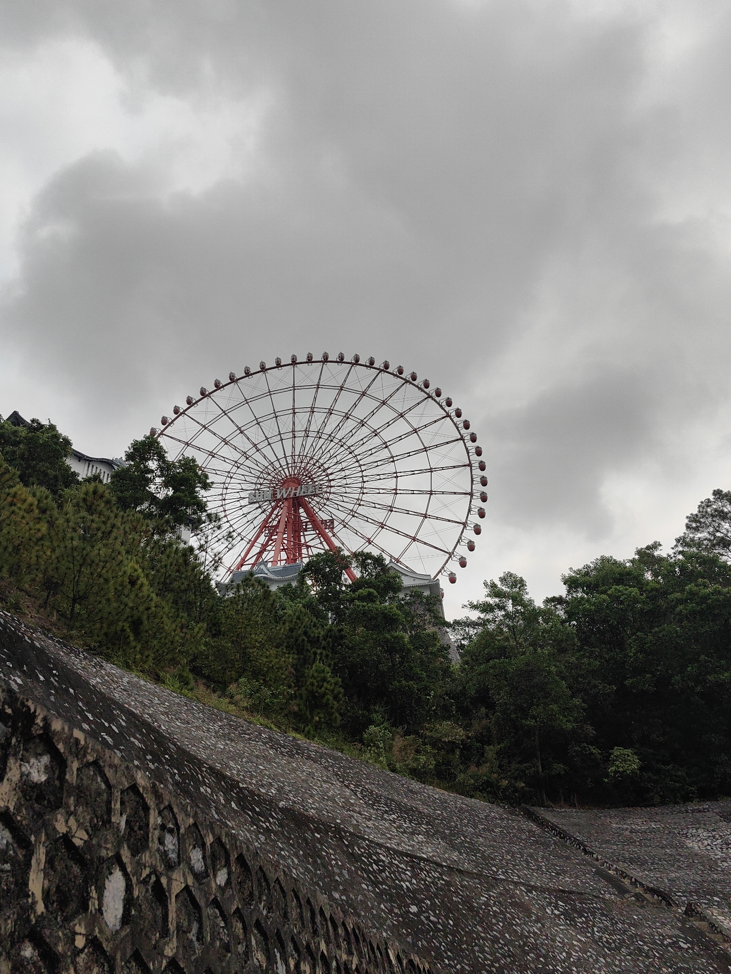 The image shows a large red and white Ferris wheel in Hong Gai Ward, Vietnam, partially obscured by trees. The Ferris wheel sits atop a hill. The sky is overcast. A stone retaining wall is in the foreground. There are no people visible in the image.