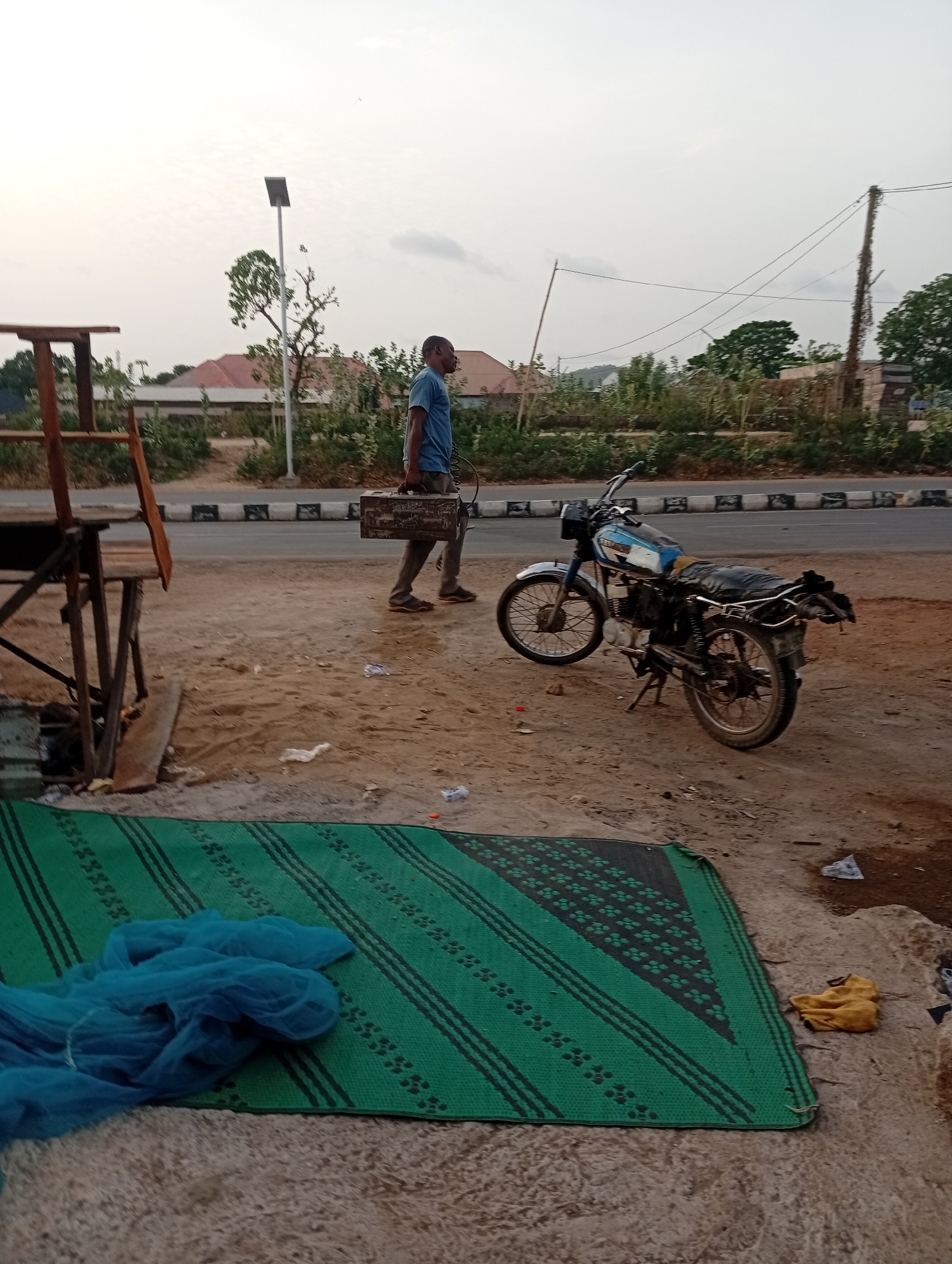 A daytime scene in Adankolo, Nigeria shows a man carrying a metal box next to a road with a black and white curb. A blue motorcycle is parked on the dirt shoulder. A green and black woven mat is on the ground near some blue netting and a yellow cloth. A crude wooden structure is on the left. A solar-powered street light is visible in the background, along with trees and low buildings. Electrical wires are strung between poles in the distance. Litter is scattered on the ground.
