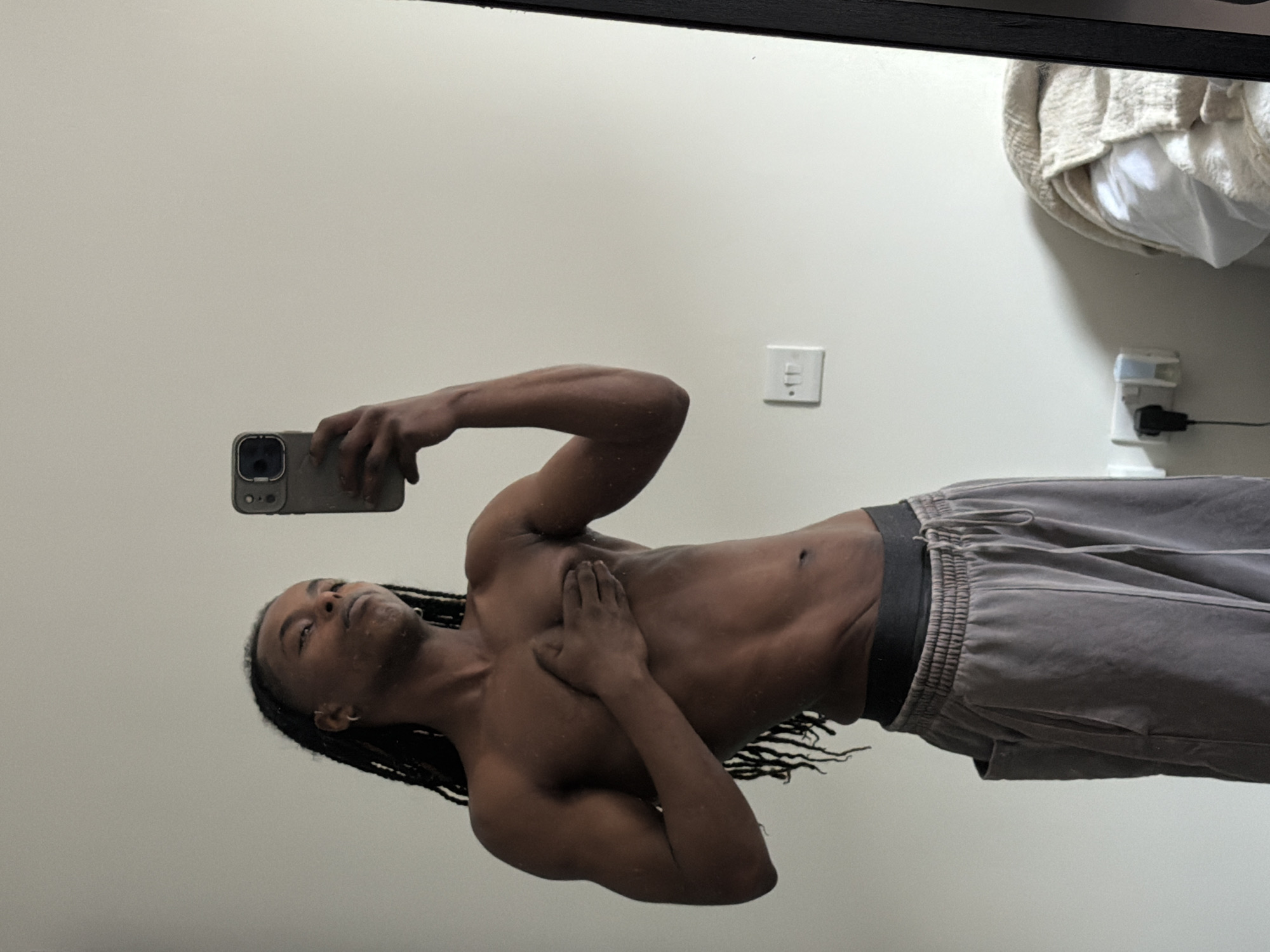 The image is a mirror selfie featuring a person with dark skin and long dark braids, appearing to be male. The subject is shirtless, displaying a toned physique, and wears grey sweatpants with an elasticated waistband and drawstring. They hold a smartphone (resembling an iPhone with a triple camera system) in their right hand to take the photo, while their left hand rests on their chest. Water droplets are visible on their skin and hair, suggesting they may have just showered or been in contact with water. The subject looks upwards and slightly to the side, towards the camera in the mirror, with a neutral expression.
The background, also reflected in the mirror, consists of a plain, light-colored wall, likely white or very light grey. On the wall, to the right of the subject, a standard white light switch is visible. Further to the right and lower, there is a white power adapter plugged into an outlet, with a black cord extending from it. Above the power adapter, a textured, light-colored blanket or throw is partially visible, resting on a surface. The lighting is bright and even, suggesting an indoor setting during daytime. No specific location cues are visible within the image, but the provided context is London, United Kingdom. There is no visible text. The original image is rotated 90 degrees counter-clockwise, showing the subject horizontally.