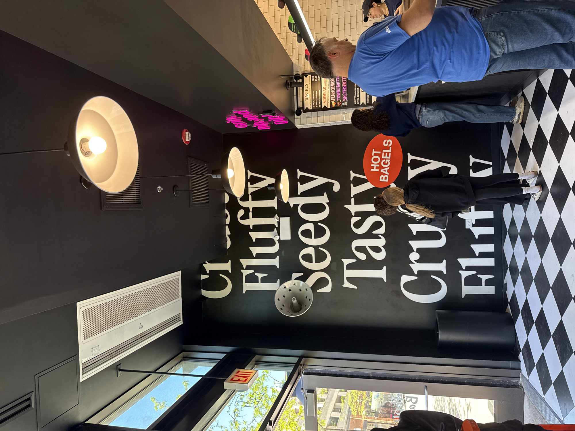 Here is a description of the image:
The image shows the interior of a bagel shop, likely in an urban setting. The focus is on a black wall with large, stylized words in white: "Crispy," "Fluffy," "Seedy," "Tasty," "Crusty," and "Flirt," arranged vertically. A red circle with the words "HOT BAGELS" is also visible.
There are several people in the scene. A man wearing a blue shirt is standing near the counter, and two other customers are lined up, waiting to order. The counter area has a menu board listing bagels and schmears, with prices. Fluorescent lights are illuminating this space.
The flooring is a checkered black and white pattern. An air conditioning unit is mounted high on the wall, near a window and an illuminated "EXIT" sign is hanging near the entrance.
Based on textual clues such as "Monty's Dairy Free Vegan," and the style of the bagel shop, it is possible the location is in Chicago, United States of America.