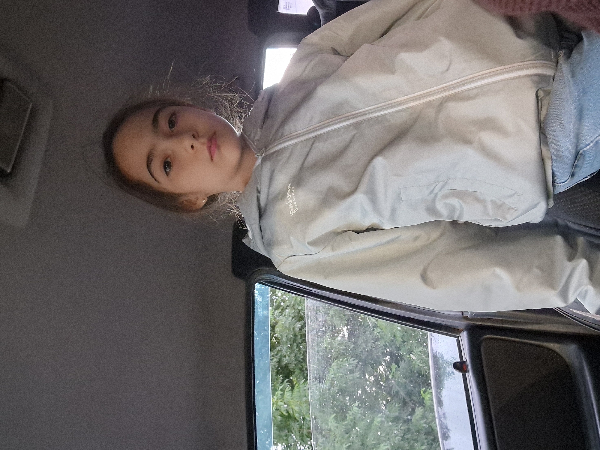 In the backseat of a car, a young girl with brown hair and brown eyes is photographed from the chest up. She is wearing a light grey hooded jacket with a zipper and blue jeans. The car interior is dark grey, with a black door panel visible to the left and a window showing green trees outside. The scene appears to be a casual snapshot, possibly taken during a drive. The location is identified as Kursavka, Russia.