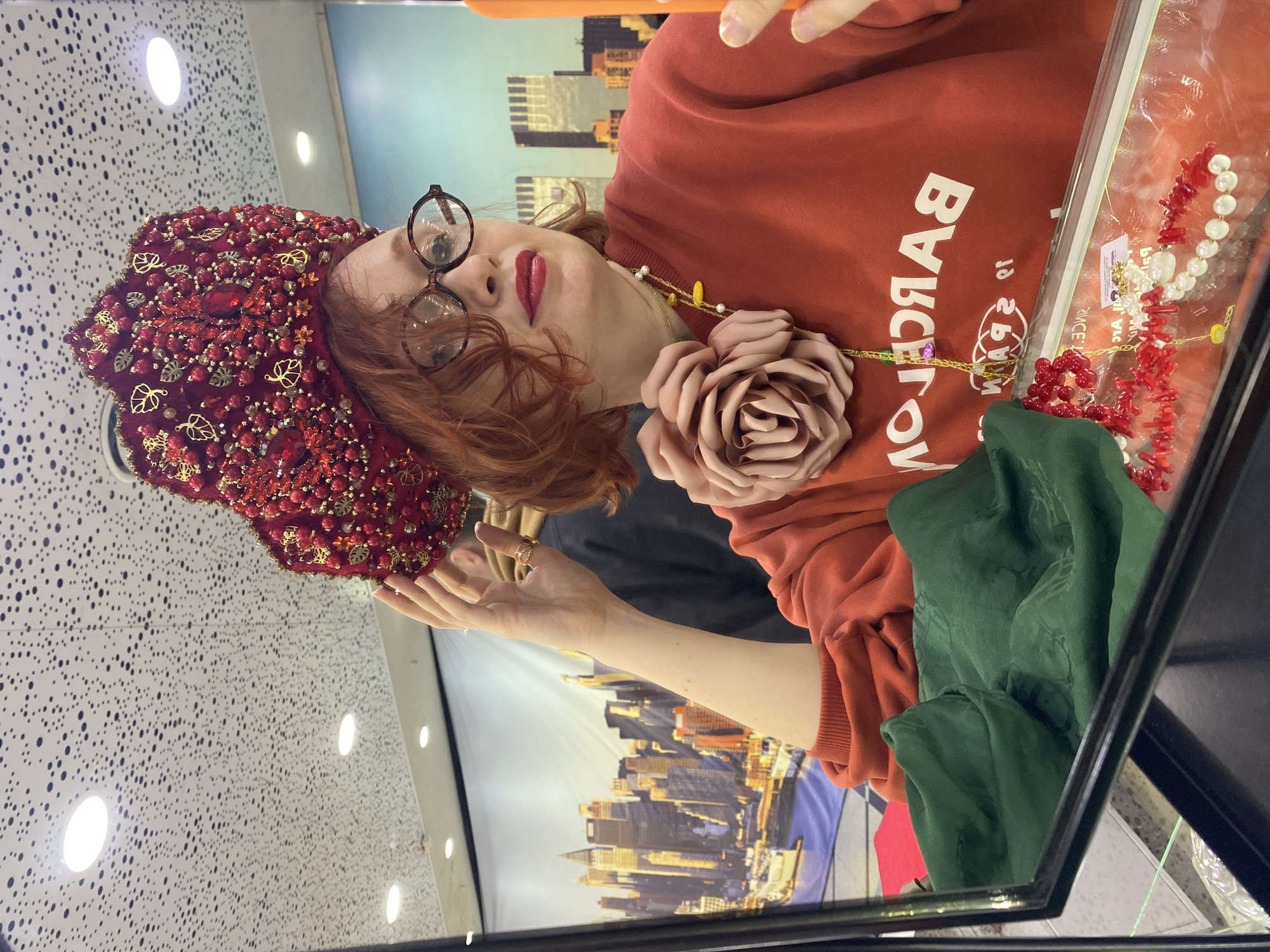 A woman is reflected in a glass case, likely a display in a store. She has reddish-auburn hair, wears round glasses, red lipstick, and a red patterned head covering with decorative stones. She is wearing an orange-red sweatshirt with the word "BARCELONA" printed vertically. The sweatshirt is adorned with a large fabric rose and a chain necklace.
In the glass case, jewelry, including a necklace with red coral and white pearls, is visible. A crumpled green fabric is also present in the case. The background shows a cityscape of tall buildings, possibly printed on a wall or poster.
Based on the location metadata, this photograph was taken in Yekaterinburg, Russia.