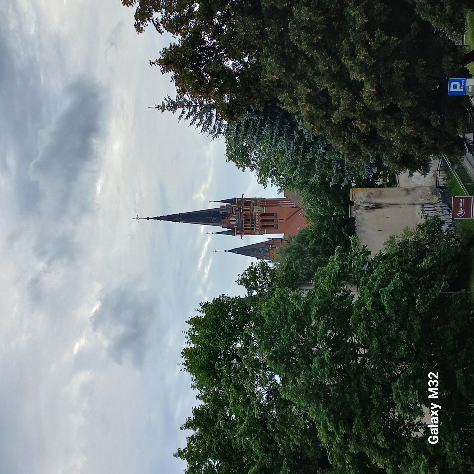 A medium shot captures the spires of a red brick church against a cloudy sky, framed by dense green trees. The church features a tall, dark spire topped with a cross, and a shorter, similarly shaped spire to its left. A clock face is visible on the main spire. In the foreground, the lower branches and leaves of lush trees obscure the base of the church and a light-colored building with graffiti on its lower section. A sign with a blue "P" for parking is visible near the bottom right corner. The "Galaxy M32" watermark is present in the bottom left corner. This scene is likely within the city of Olsztyn, Poland, given the camera watermark, though specific landmarks are not definitively identifiable from the image alone.