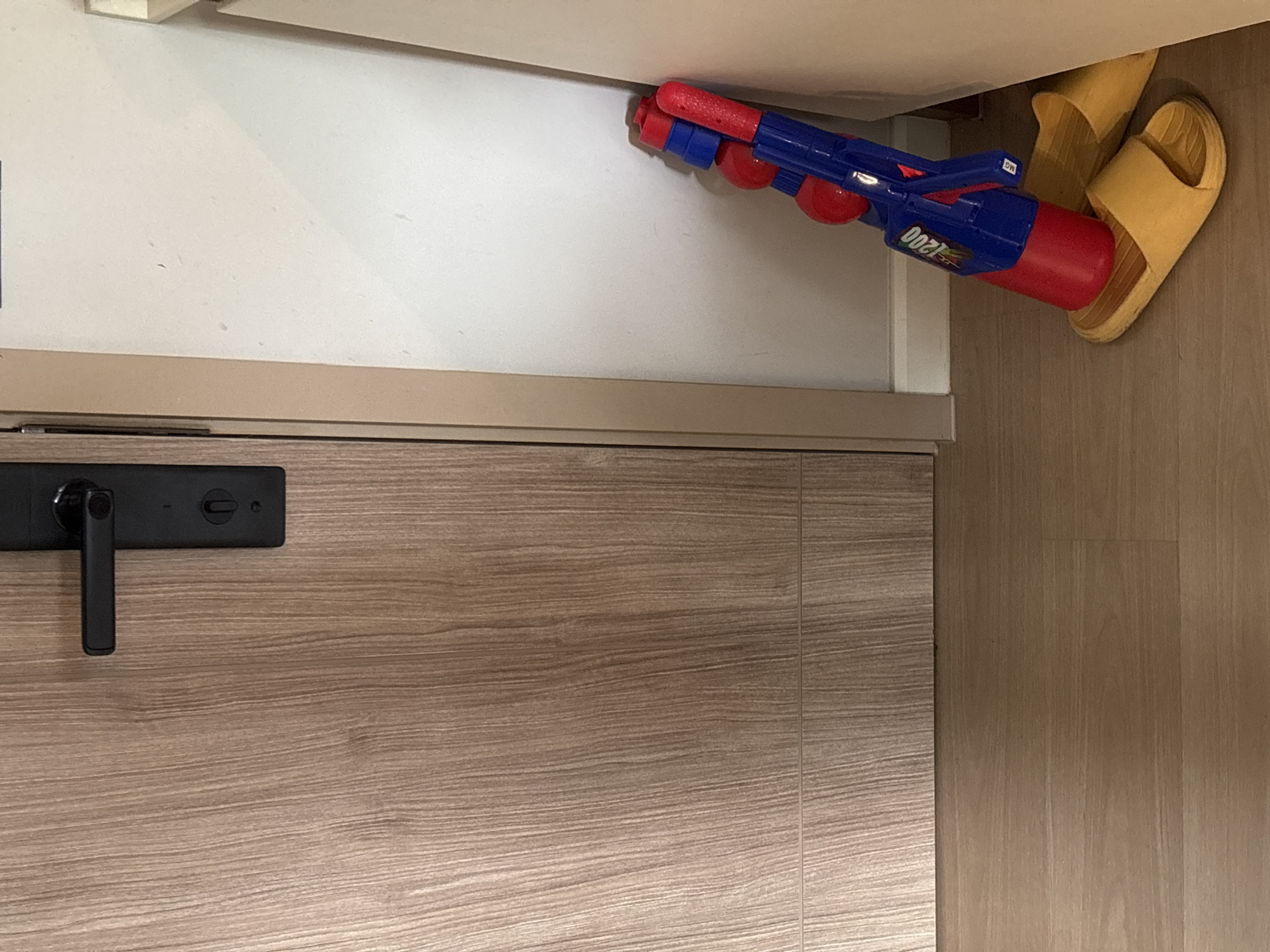 The image shows a partially open cabinet or storage area from an elevated perspective. Inside, a blue and red water gun and a pair of yellow sandals are visible. The background includes the cabinet door, which has a horizontal wood grain pattern and a black door handle. The adjacent wall is a light, solid color. The location is not discernible from the image content alone. Without further information, determining if this image is from Choeng Thale, Thailand is not possible.
