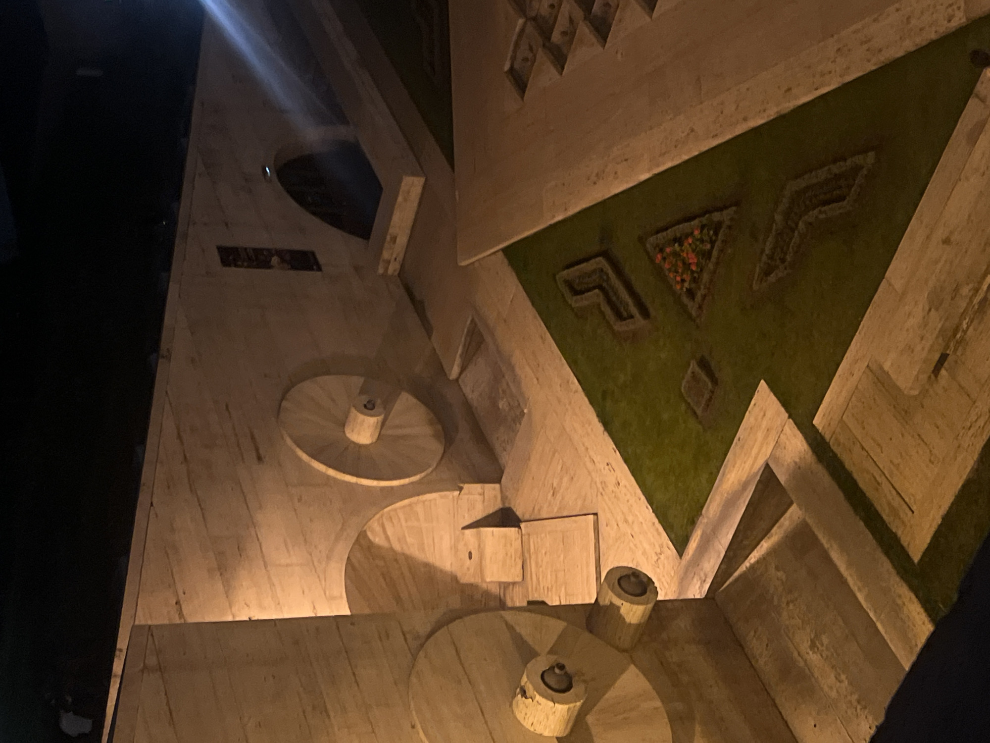 This media file captures an illuminated architectural complex at night, likely the Cascade in Yerevan, Armenia. The scene is dominated by multi-level structures made of light-colored, possibly travertine, stone. Geometric patterns and shapes are prominent, including walls, steps, and inclined surfaces.
Several circular platforms, possibly bases for fountains or sculptures, are visible, each with a central cylindrical element. Interspersed with the stone architecture are sections of green landscaping, including a triangular planter filled with small red/orange flowers and an arrow-shaped green area.
The complex is bathed in warm, artificial lighting, casting strong shadows and highlighting the textures and contours of the stone. A bright light source or glare is visible in the upper left, contributing to the nighttime atmosphere. The absence of people suggests a quiet moment at the location.
On one of the stone walls, a rectangular panel contains faint, illegible markings or an inscription. No specific activity or event is taking place; the image focuses on the architectural beauty and its nighttime illumination.