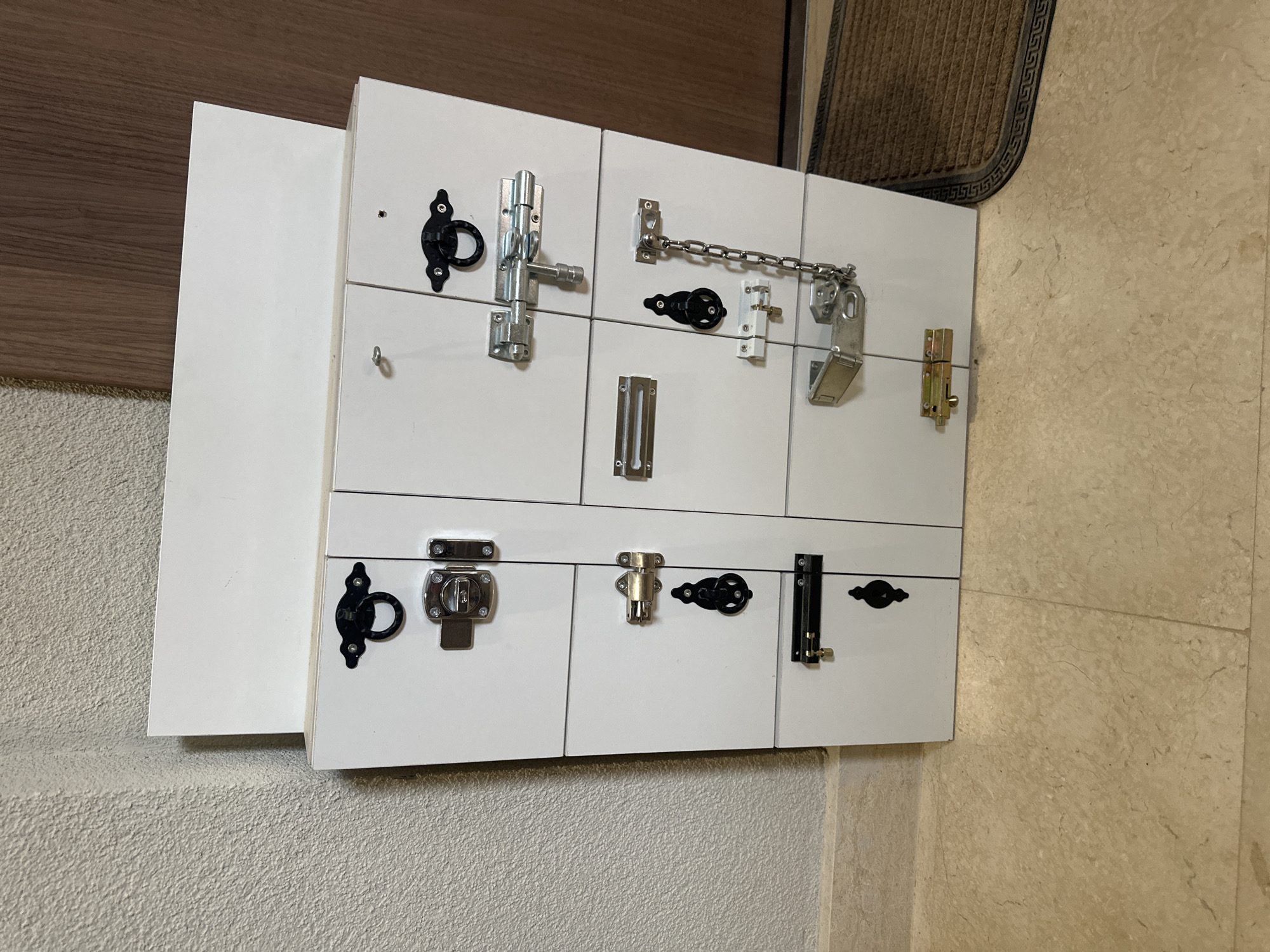 The image shows a white wooden activity board mounted on a wall in what appears to be a domestic setting. The board is arranged as a 3x3 grid and features various types of door hardware including latches, locks, hooks, chains, and bolts. The wall behind the board is painted or covered in a beige or off-white material. A dark brown mat or doorframe is visible above and to the right of the board. The location in Budva, Montenegro is not discernible from the image. No people are visible, and there are no actions or interactions occurring in the frame.