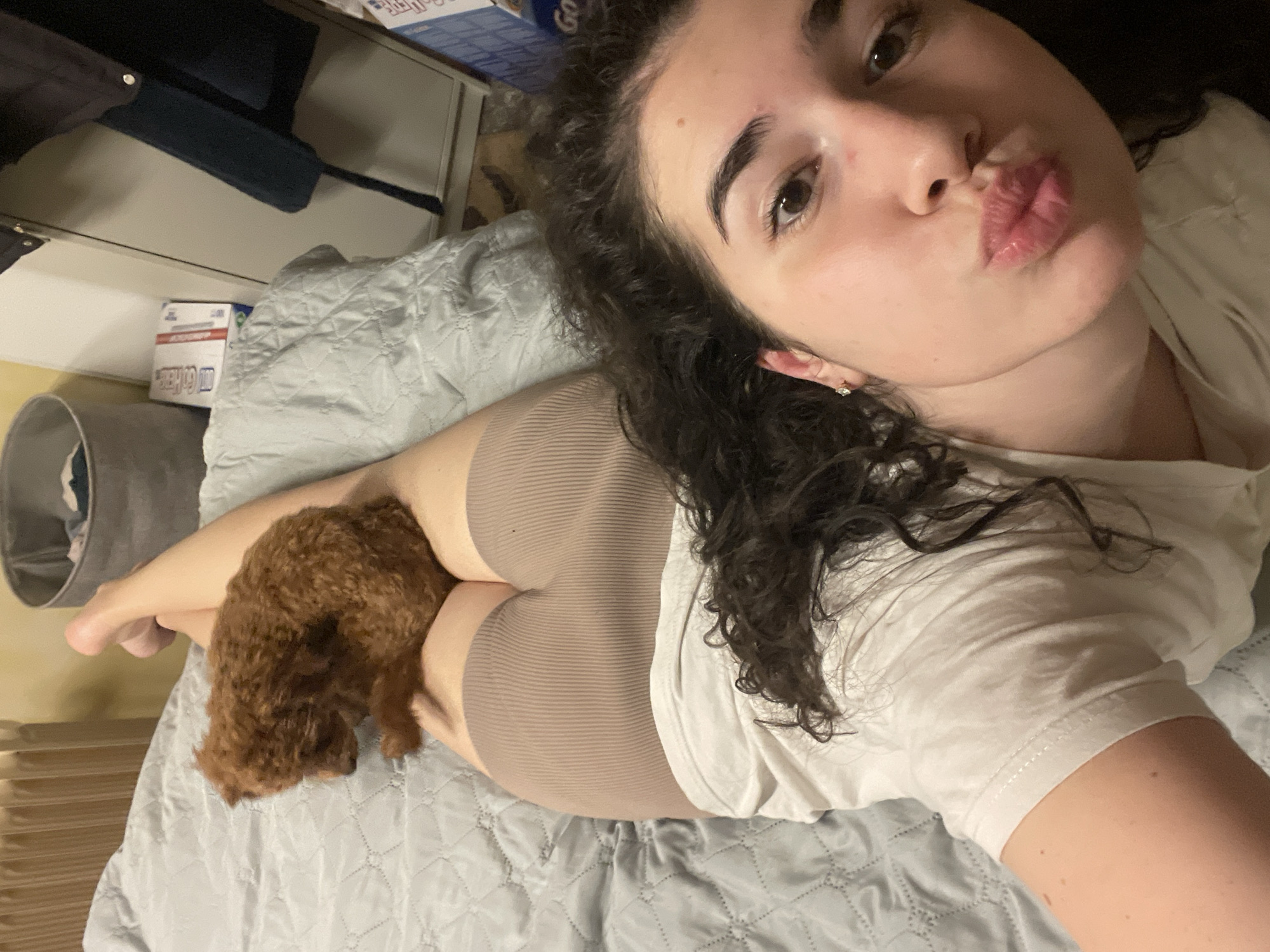 A woman is lying on a bed with a small, brown poodle beside her. She is wearing a white t-shirt and tan-colored shorts. The bed has a gray, quilted comforter. The background includes a nightstand with items on top and a gray, round object. The location is not specified, and there is no indication that it is in Sofia, Bulgaria.