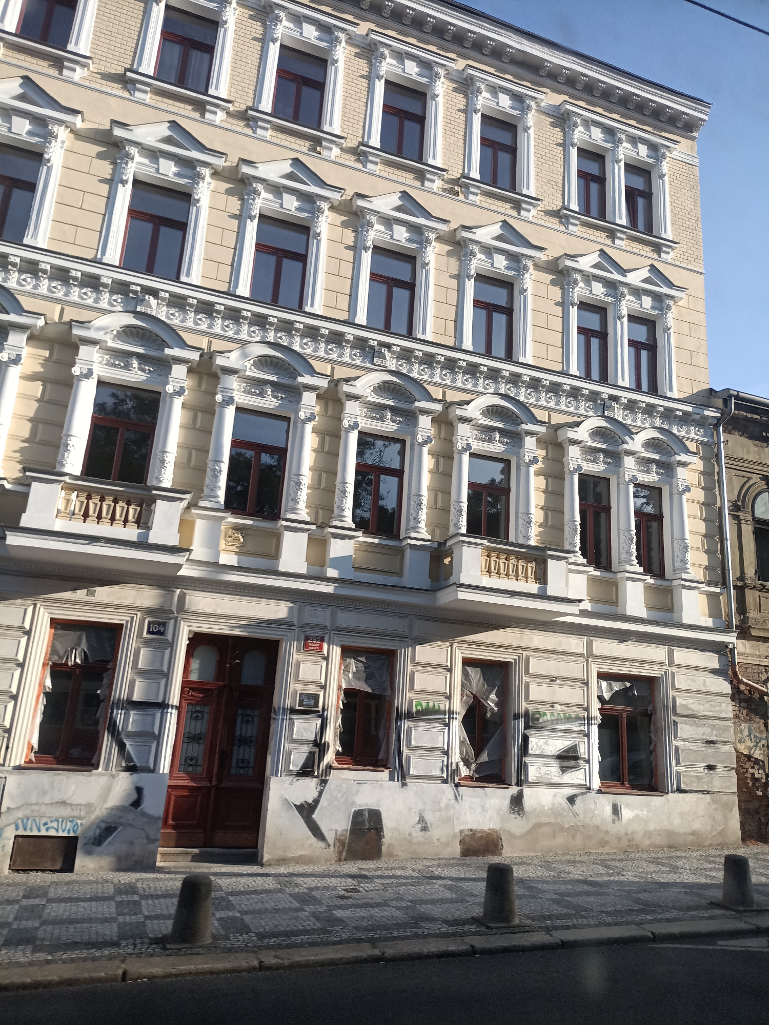 The image shows the facade of a multi-story building in Prague, Czechia. The building has a light yellow facade with ornate white decorative elements around the windows and cornices. The windows on the upper floors appear intact and reflect the blue sky. The lower floors have some damaged windows covered with plastic sheeting, and graffiti is visible on the ground floor walls. A red double door serves as the entrance. Cobblestone pavement is in front of the building, and two concrete bollards are visible on the sidewalk. An adjacent building to the right appears older and in a state of disrepair. The scene is illuminated by sunlight, casting shadows on the pavement.